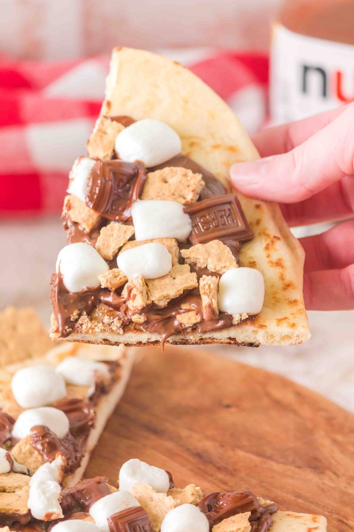 A hand holds a slice of dessert pizza topped with chocolate, graham crackers, and marshmallows.