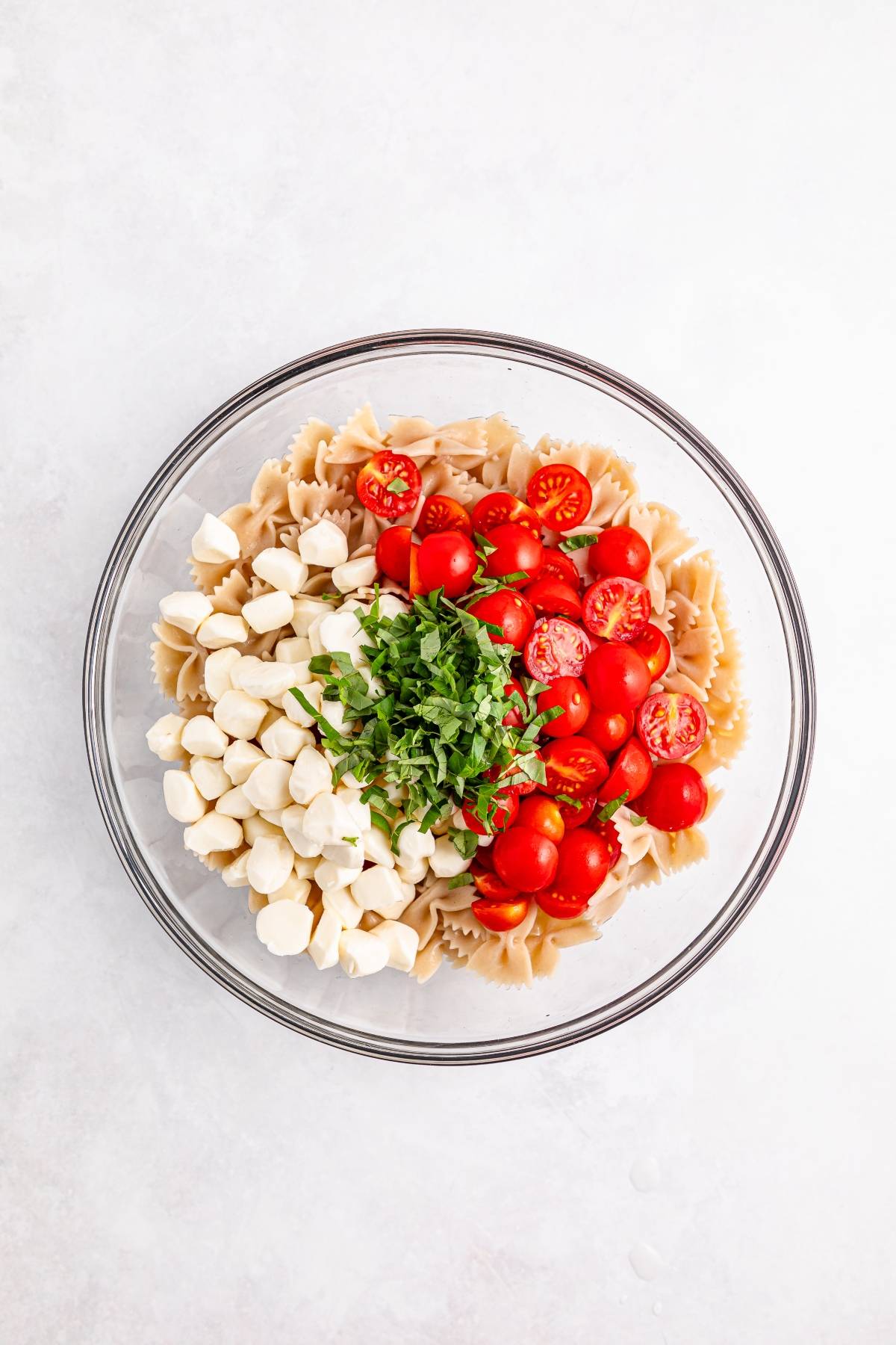 A glass bowl with bowtie pasta, cherry tomatoes, mozzarella balls, and chopped basil on a white surface.