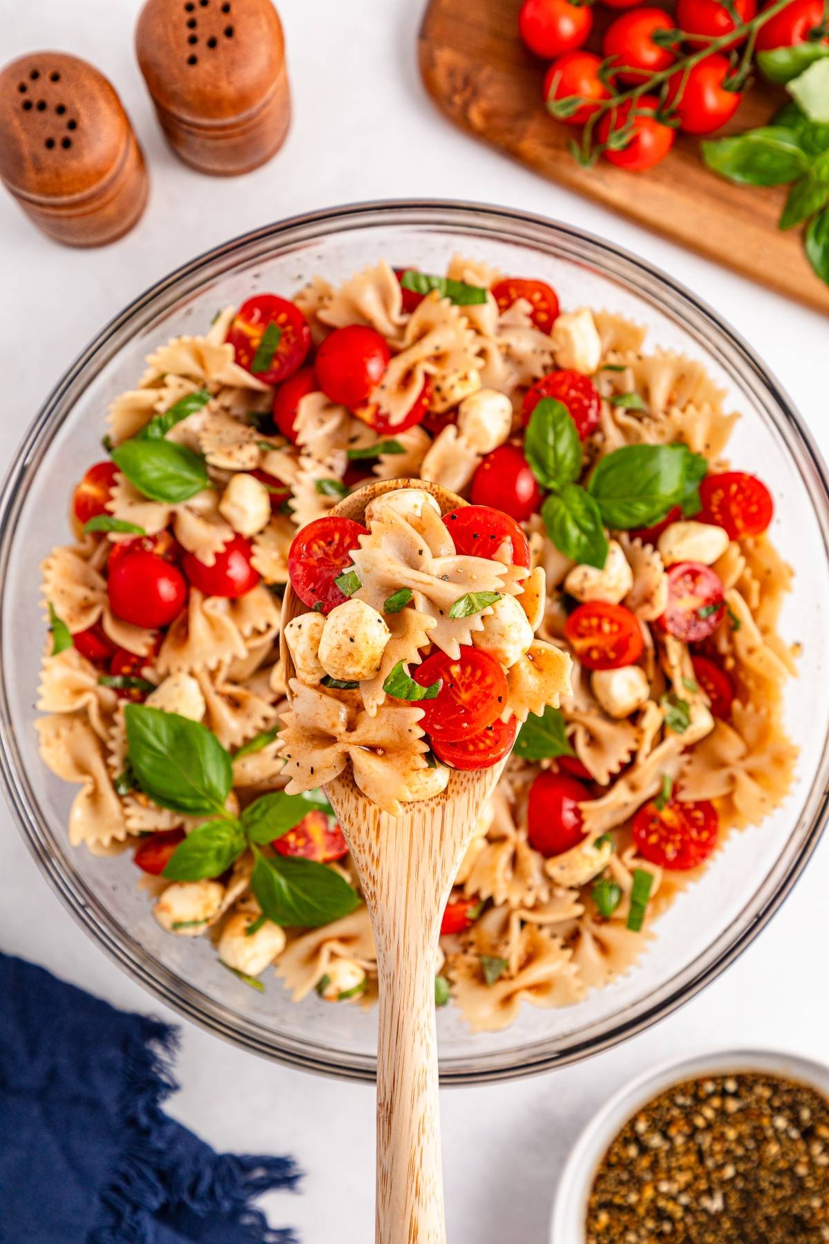 A wooden spoon holds bowtie pasta salad with cherry tomatoes, mozzarella, and basil over a glass bowl.