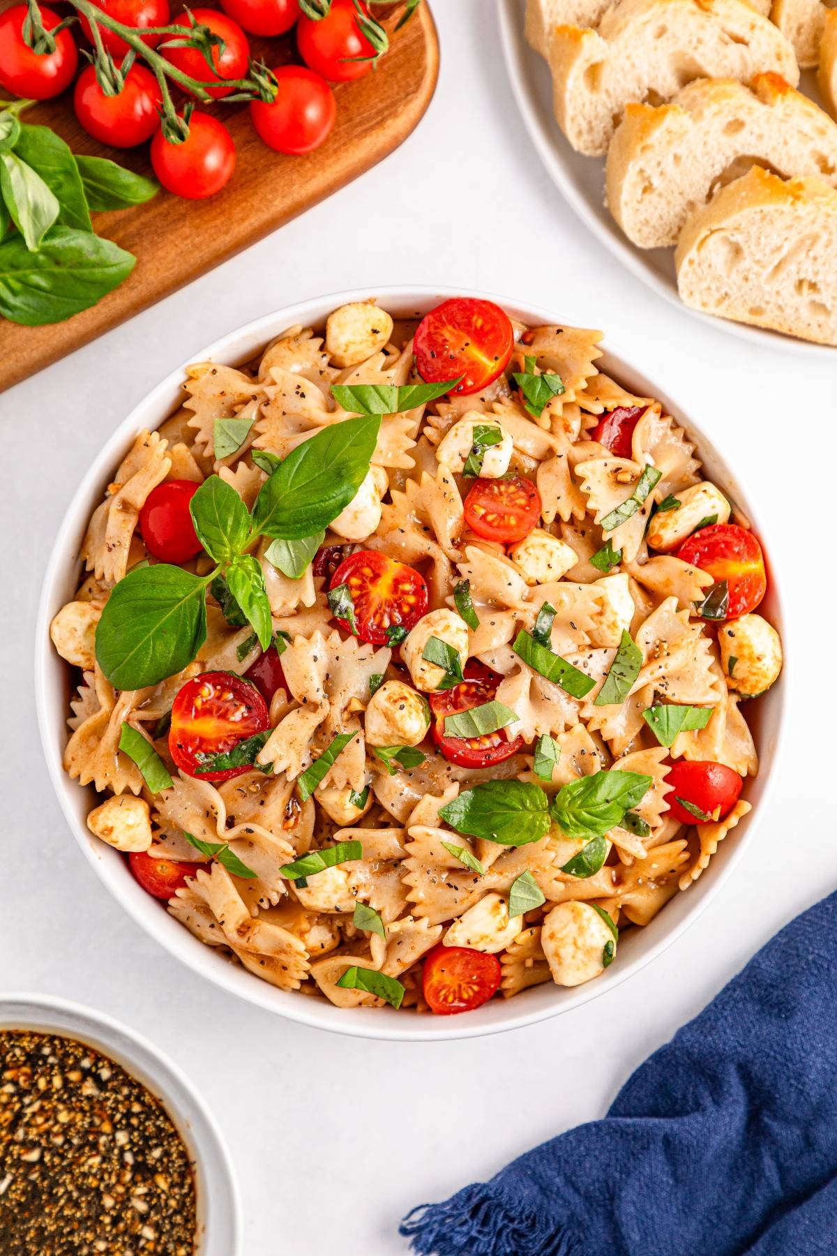 Bowl of bowtie pasta salad with cherry tomatoes, mozzarella, basil, and herbs, next to bread and tomatoes.