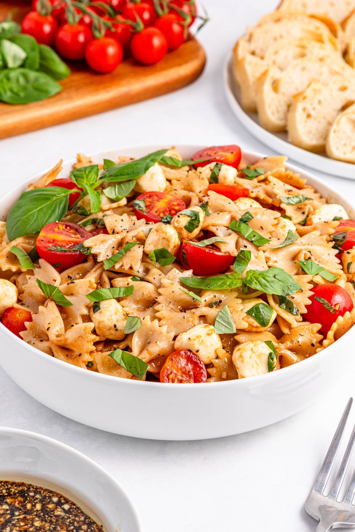 A bowl of bowtie pasta salad with cherry tomatoes, mozzarella, basil, and black pepper, surrounded by bread and tomatoes.