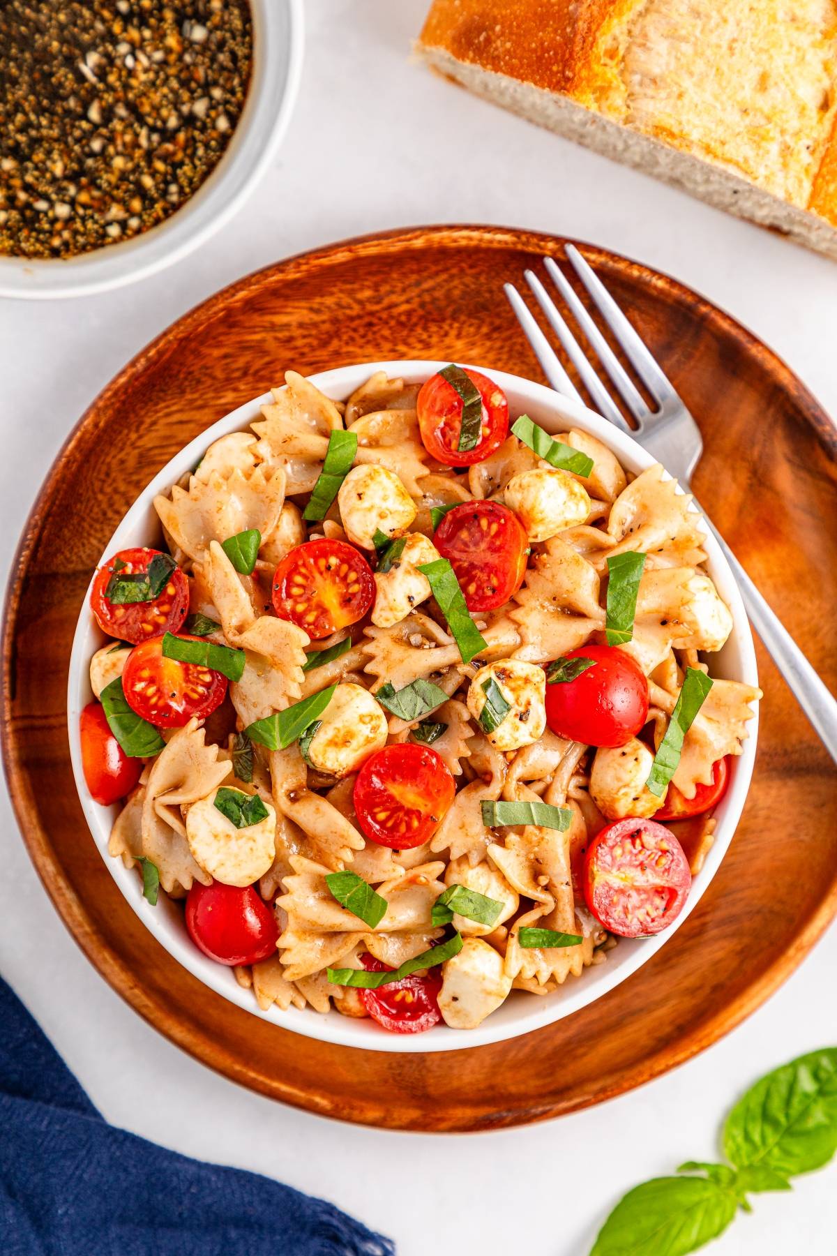 Bowl of pasta salad with cherry tomatoes, mozzarella, and basil on a wooden plate, with bread and dressing nearby.