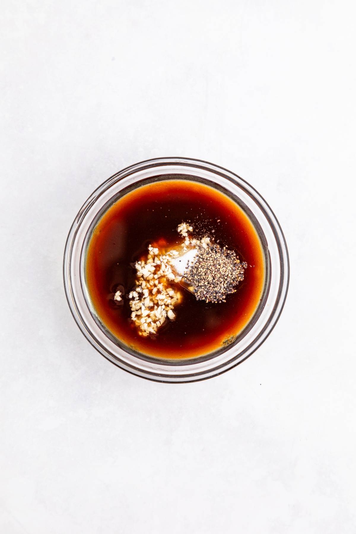 A glass bowl with dark sauce, minced garlic, and cracked black pepper on a white surface.