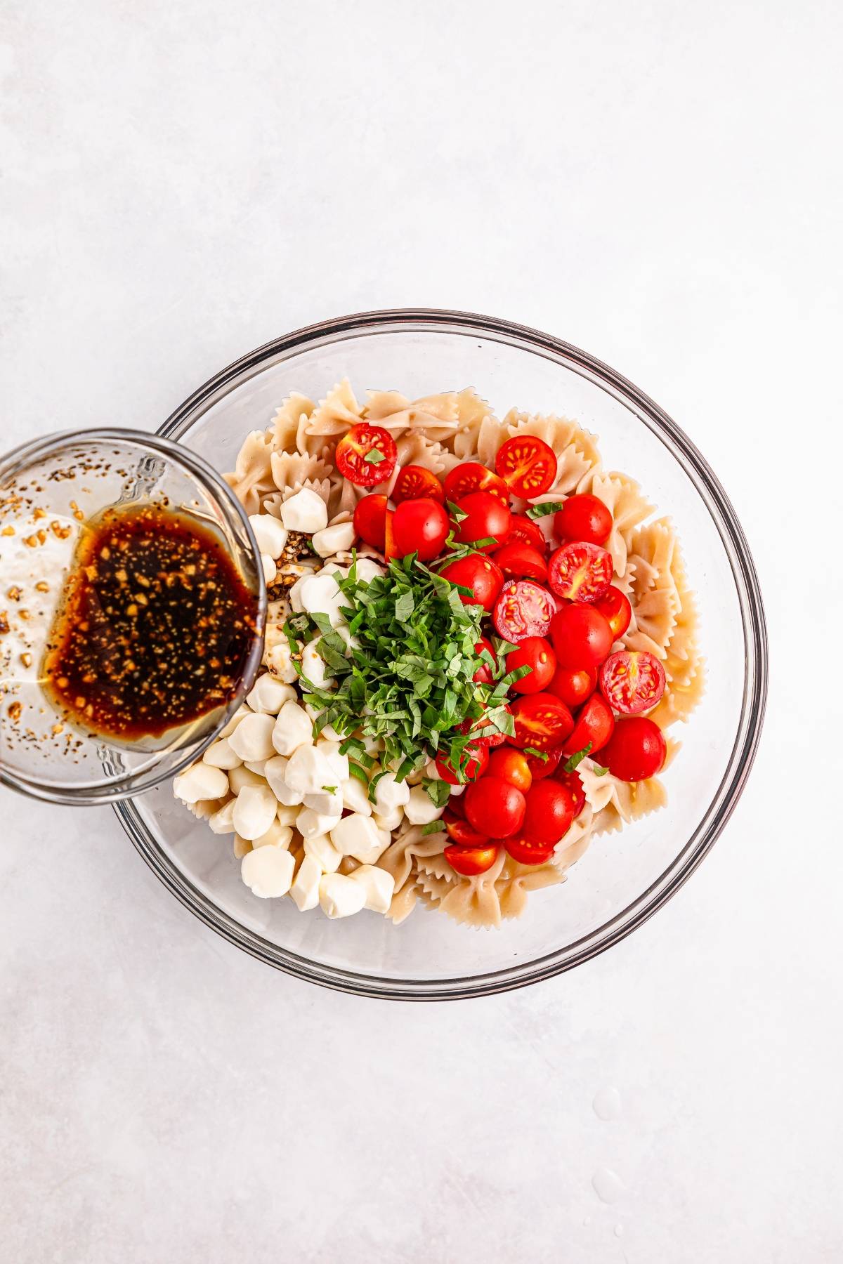 Glass bowl with bowtie pasta, cherry tomatoes, mozzarella, basil, and a cup of vinaigrette on the side.