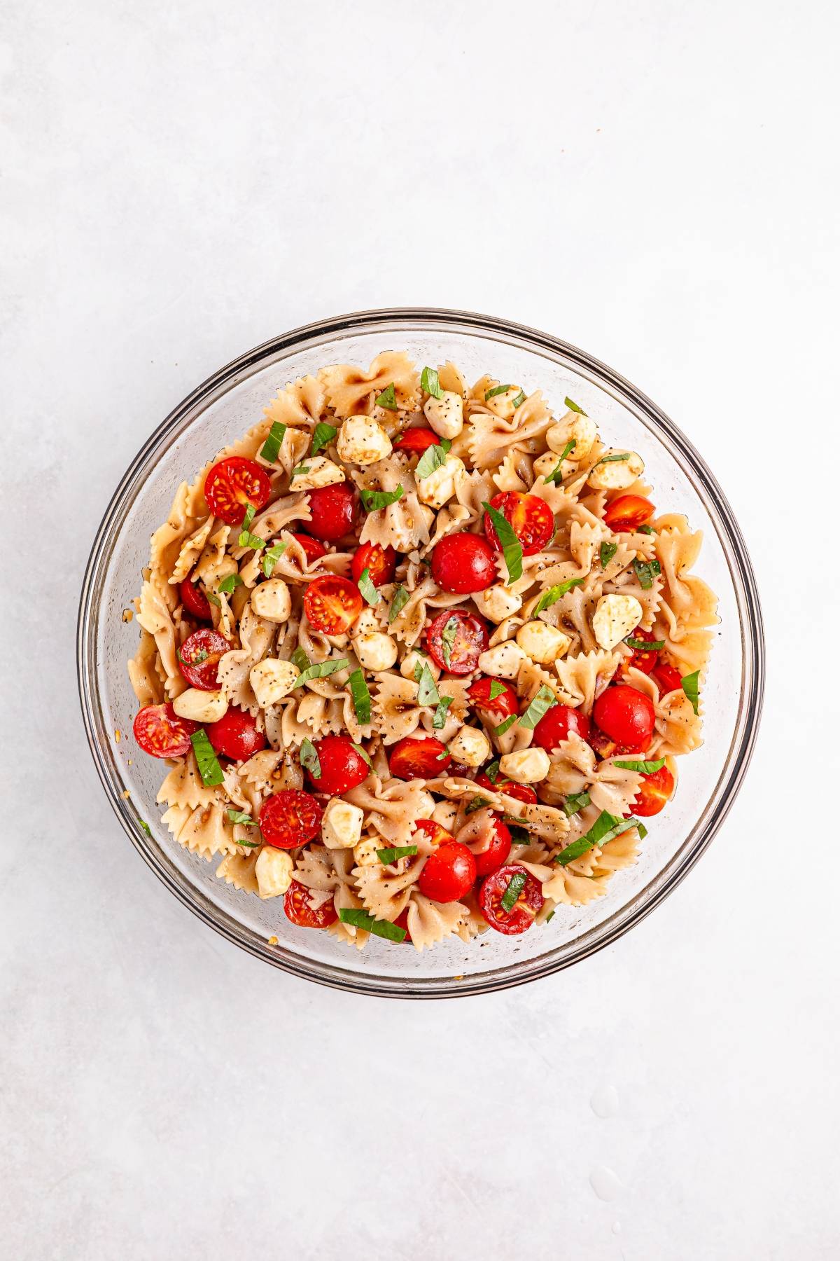 Glass bowl of pasta salad with cherry tomatoes, mozzarella, basil, and Italian seasoning on a white background.