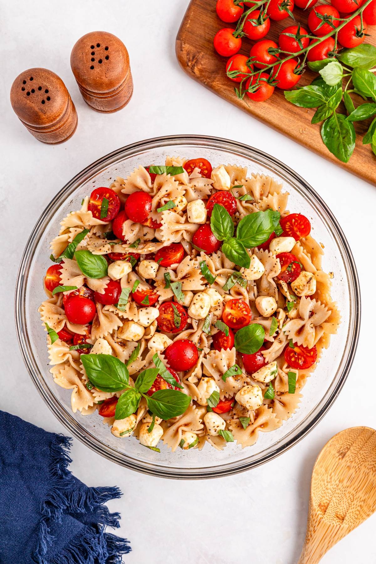A bowl of bow tie pasta salad with cherry tomatoes, mozzarella, basil, and herbs on a white table.