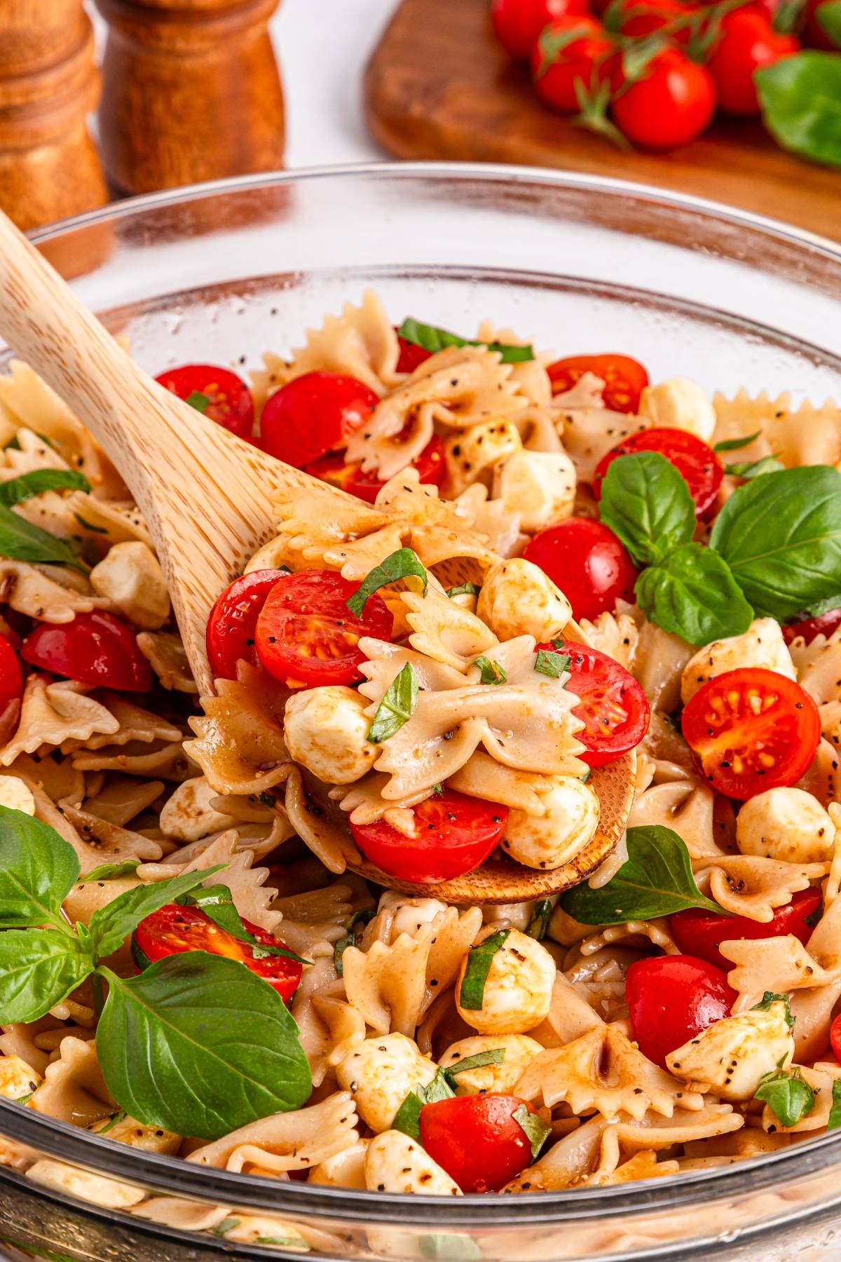 A wooden spoon scoops bowtie pasta salad with cherry tomatoes, mozzarella, and fresh basil in a glass bowl.