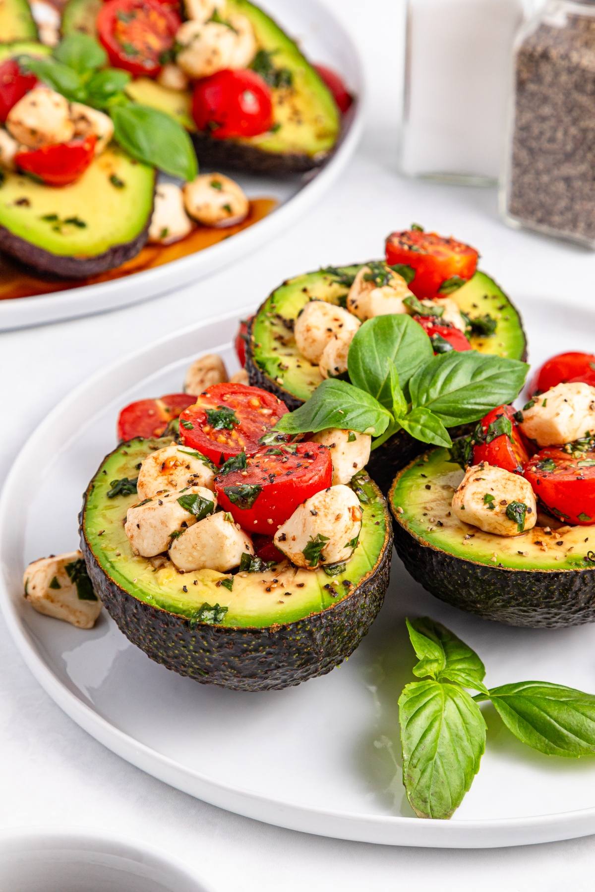 Halved avocados filled with tomatoes, mozzarella, and herbs, garnished with fresh basil on a white plate.