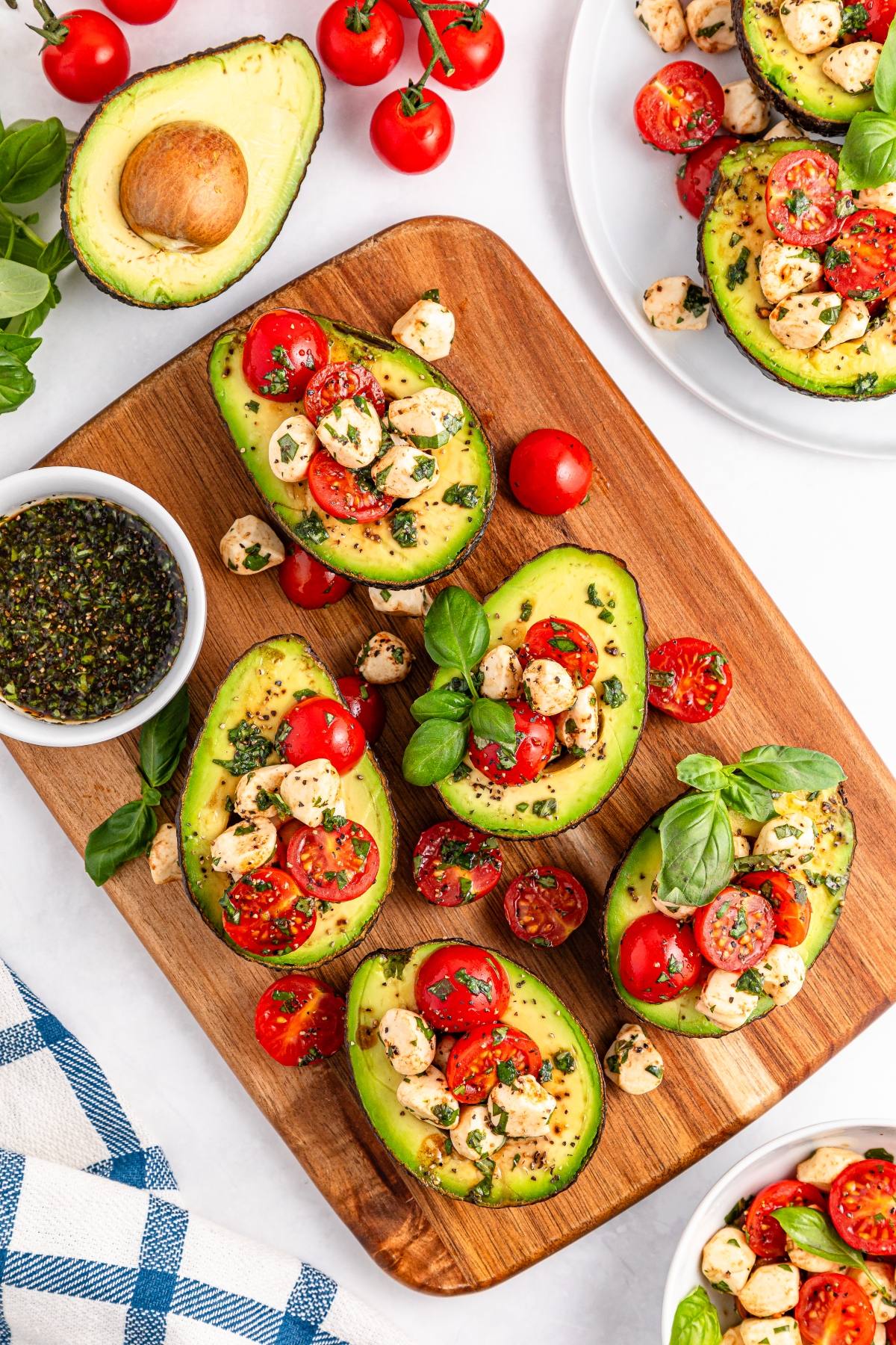 Avocado halves topped with cherry tomatoes, mozzarella balls, basil, and pesto on a wooden board.