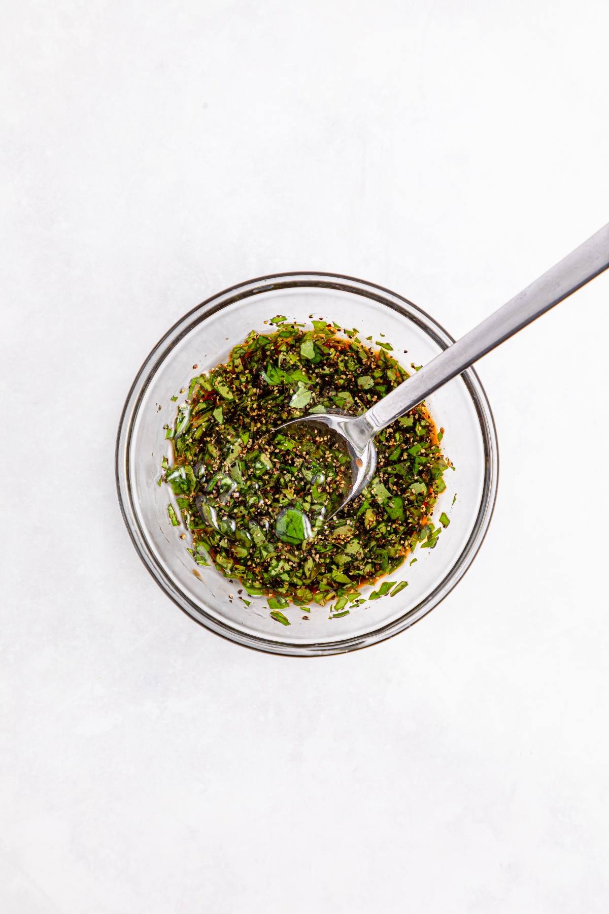 A glass bowl with chopped herbs and spices mixture, and a spoon inside, on a white background.
