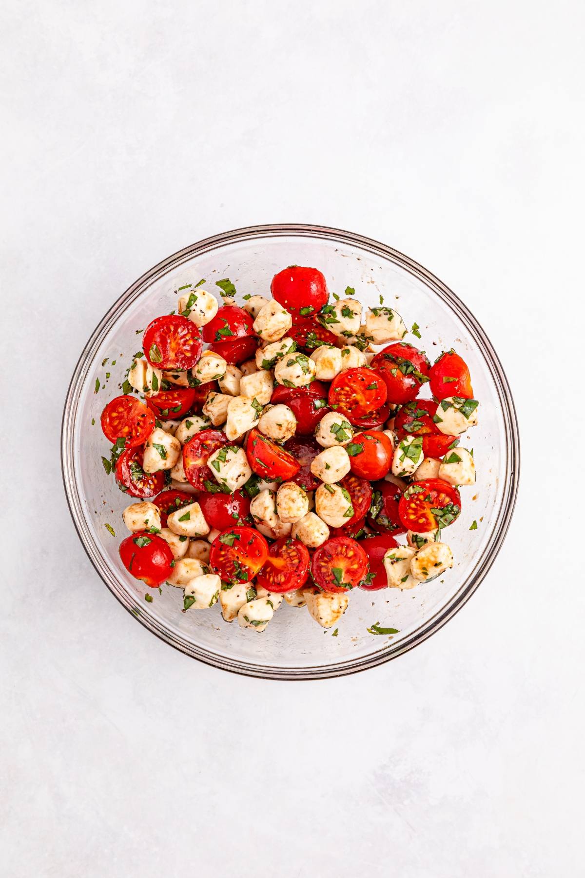 A glass bowl filled with tomato, mozzarella balls, and herbs salad on a white background.
