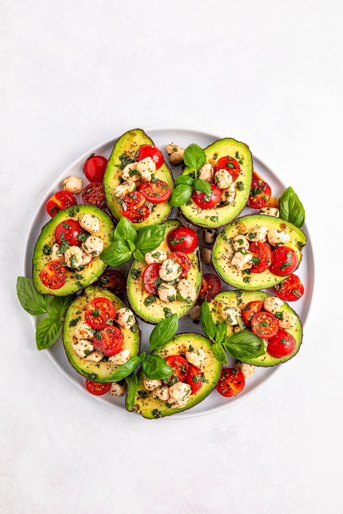 Halved avocados filled with cherry tomatoes, mozzarella balls, and herbs on a white plate, garnished with basil.