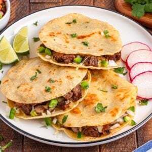Three beef quesadillas on a plate with lime wedges, sliced radishes, and garnished with fresh cilantro&mdash;these carne asada mulitas are packed with flavor and perfect for sharing.
