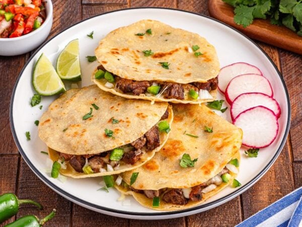 Three beef quesadillas on a plate with lime wedges, sliced radishes, and garnished with fresh cilantro—these carne asada mulitas are packed with flavor and perfect for sharing.