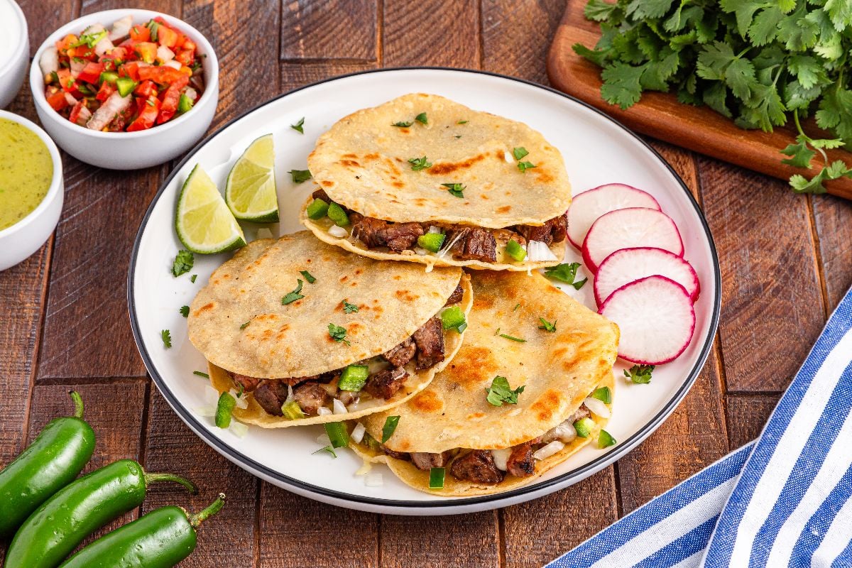 Three beef quesadillas on a plate with lime wedges, radish slices, salsa, and fresh cilantro on the side&mdash;perfect for fans of classic carne asada mulitas flavors.