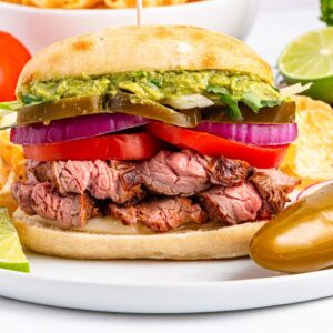 A carne asada torta steak sandwich with guacamole, onions, tomatoes, and jalape&ntilde;os, served on a plate with lime and a jalape&ntilde;o pepper.