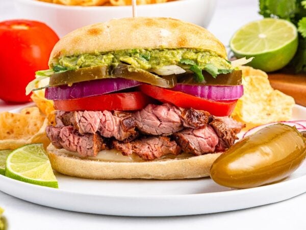 A carne asada torta steak sandwich with guacamole, onions, tomatoes, and jalapeños, served on a plate with lime and a jalapeño pepper.