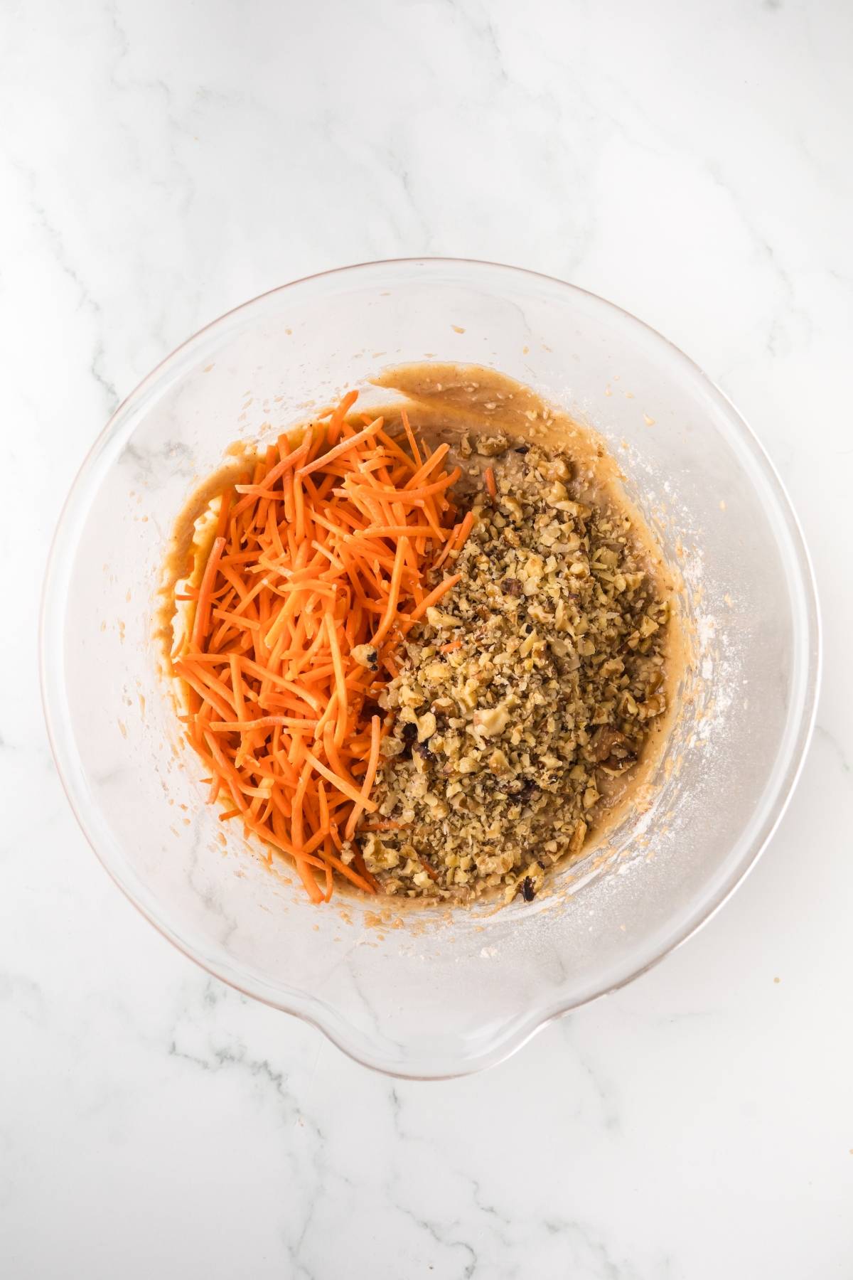 Clear glass bowl with shredded carrots, chopped walnuts, and batter on a white marble surface.