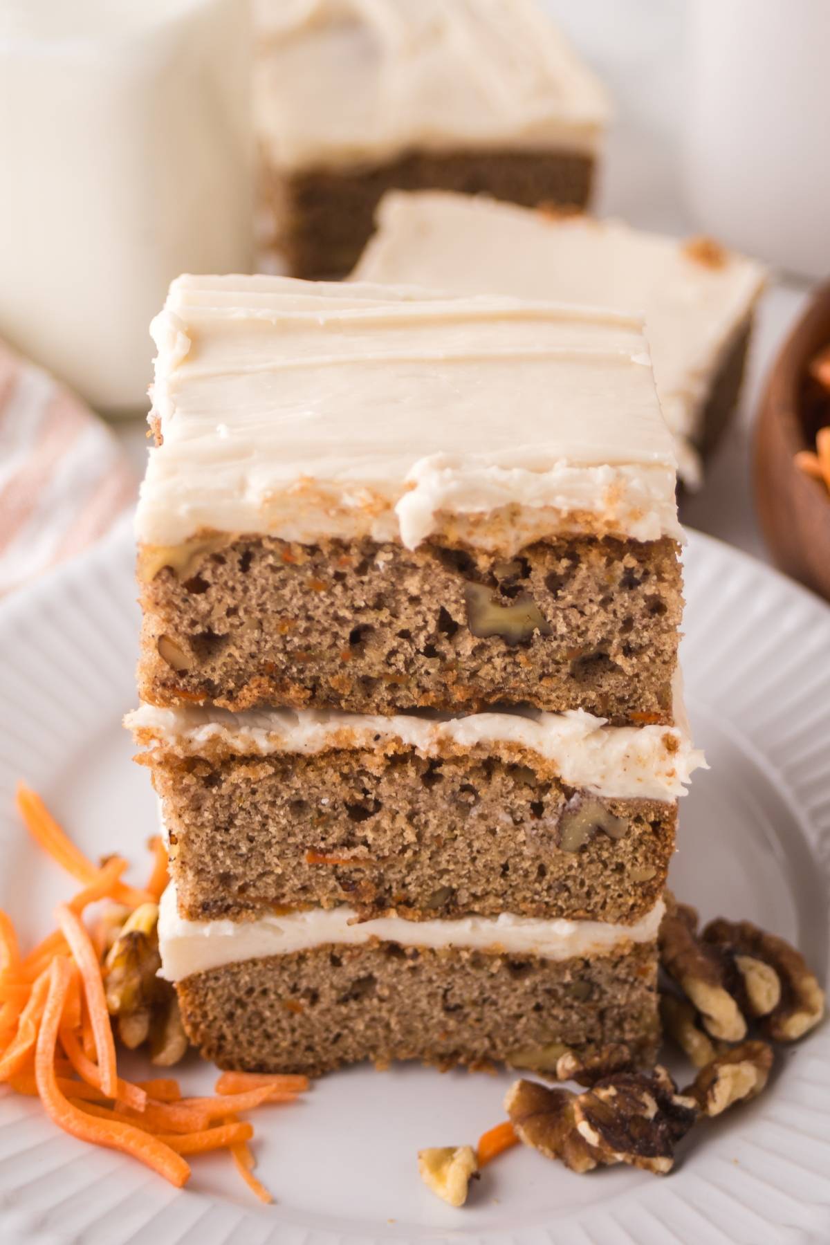 A stack of three slices of frosted carrot cake with walnuts on a white plate, garnished with carrot shreds.