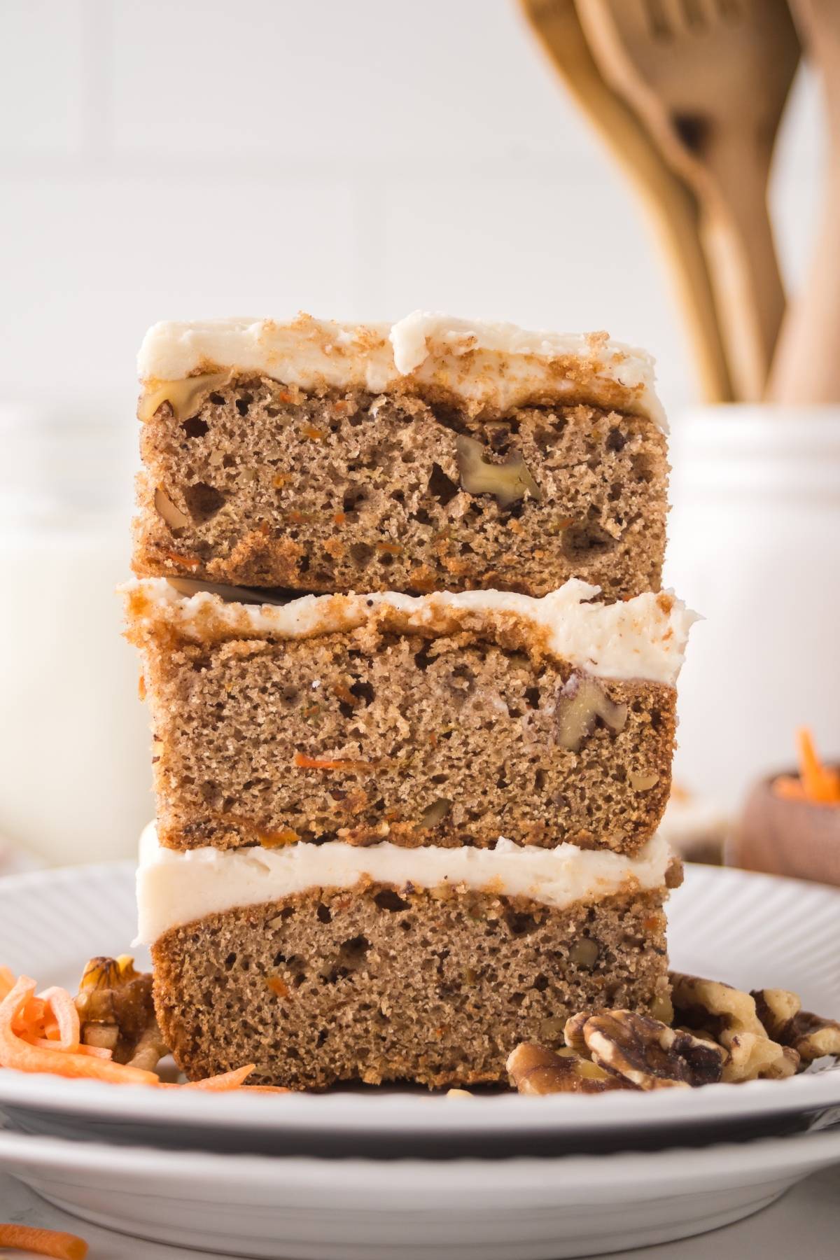 Three slices of frosted carrot cake stacked on a white plate with walnuts and shredded carrots around them.