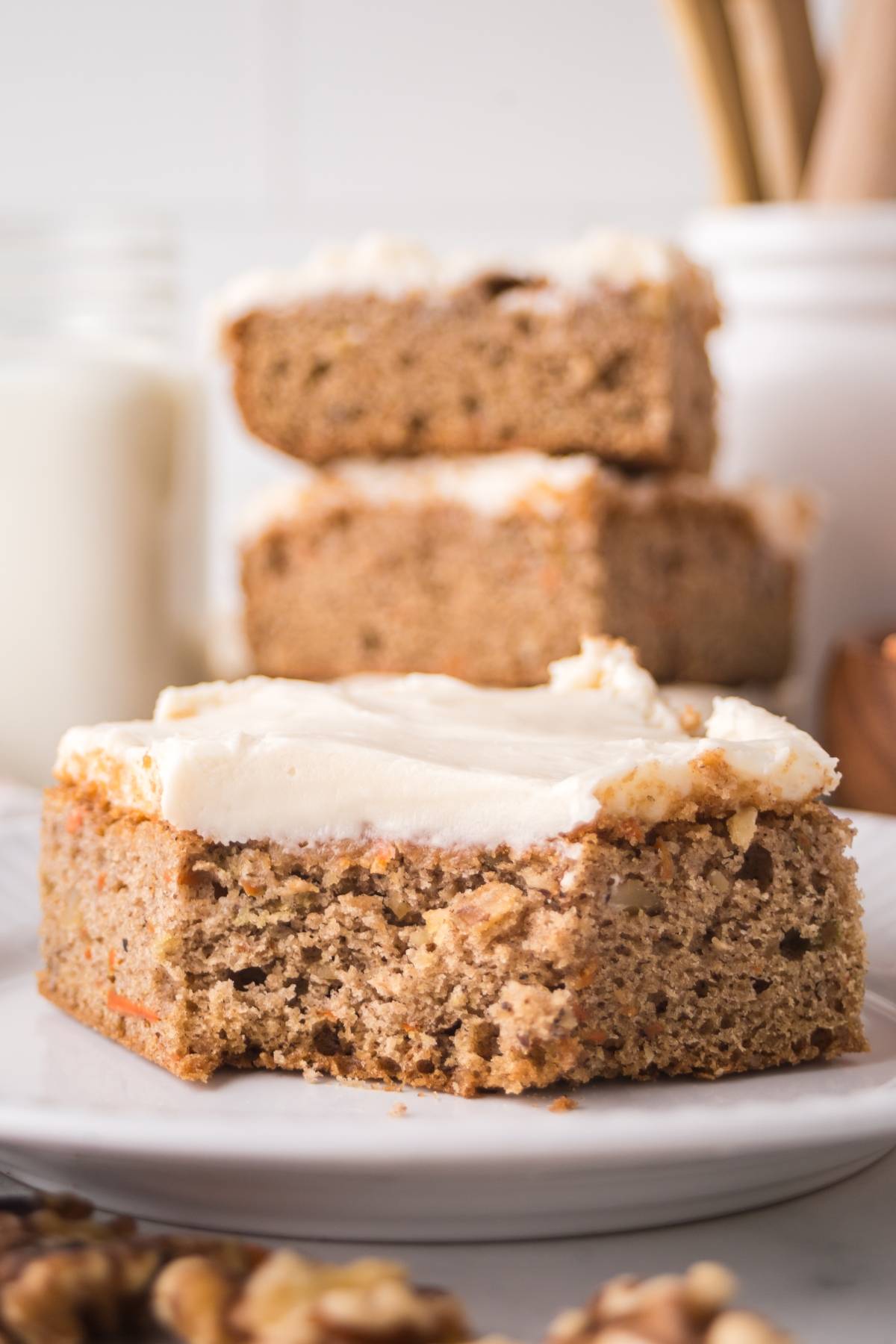 A slice of frosted banana cake with a bite taken out, stacked cake pieces and milk in the background.
