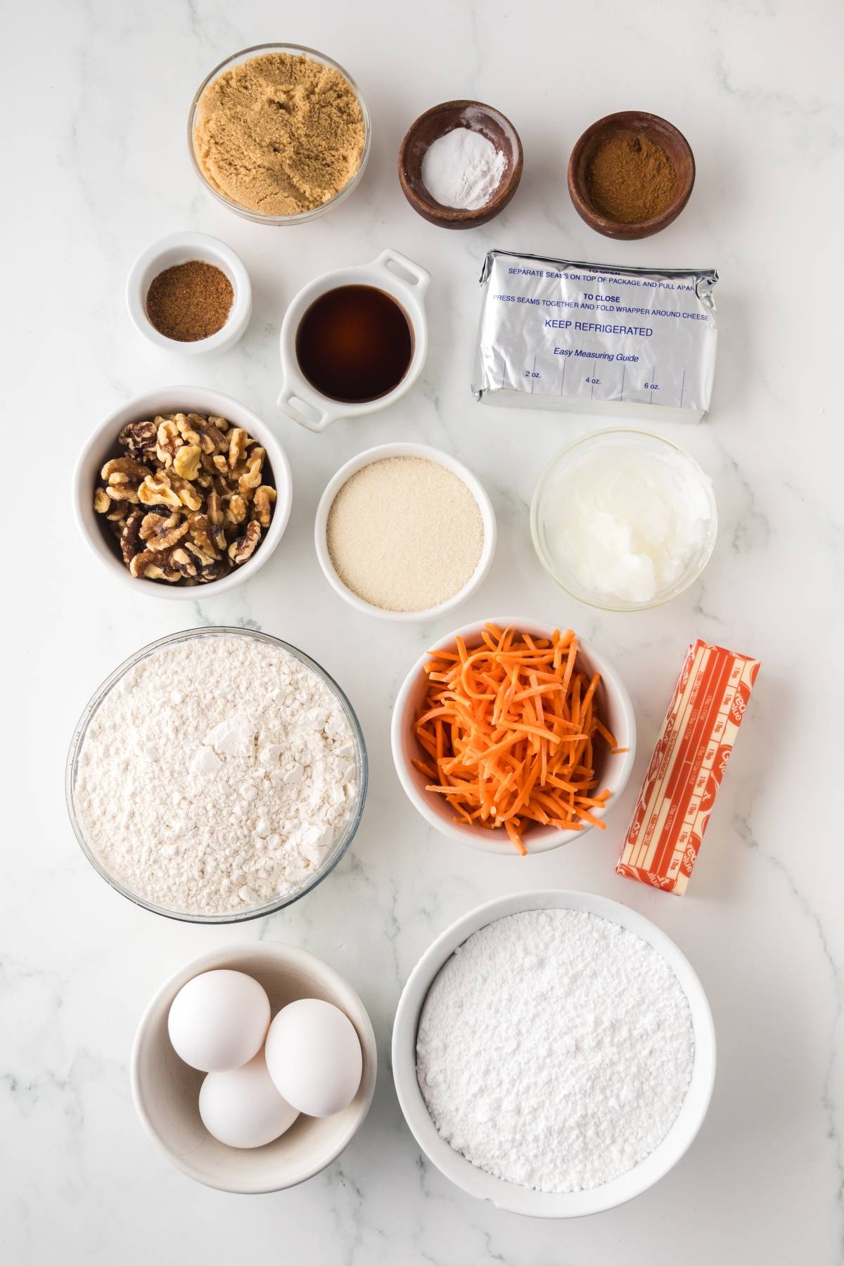 Bowls of baking ingredients including eggs, flour, sugar, walnuts, shredded carrots, butter, and spices on a white countertop.