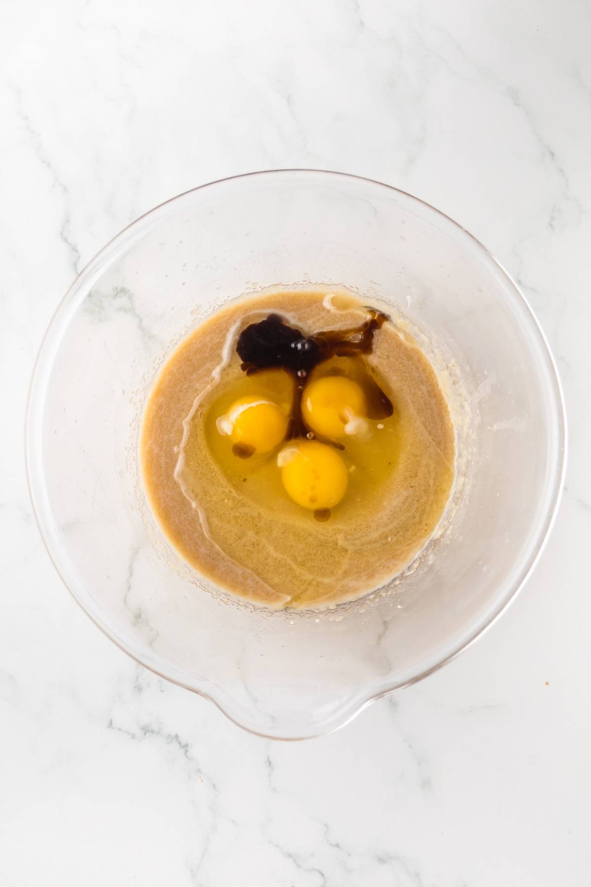 A glass bowl with three eggs, brown sugar, and vanilla extract on a white marble surface.