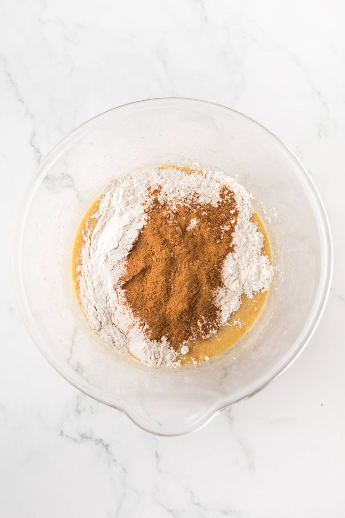 A glass bowl with flour and cinnamon on top of wet batter, ready to be mixed, on a marble surface.