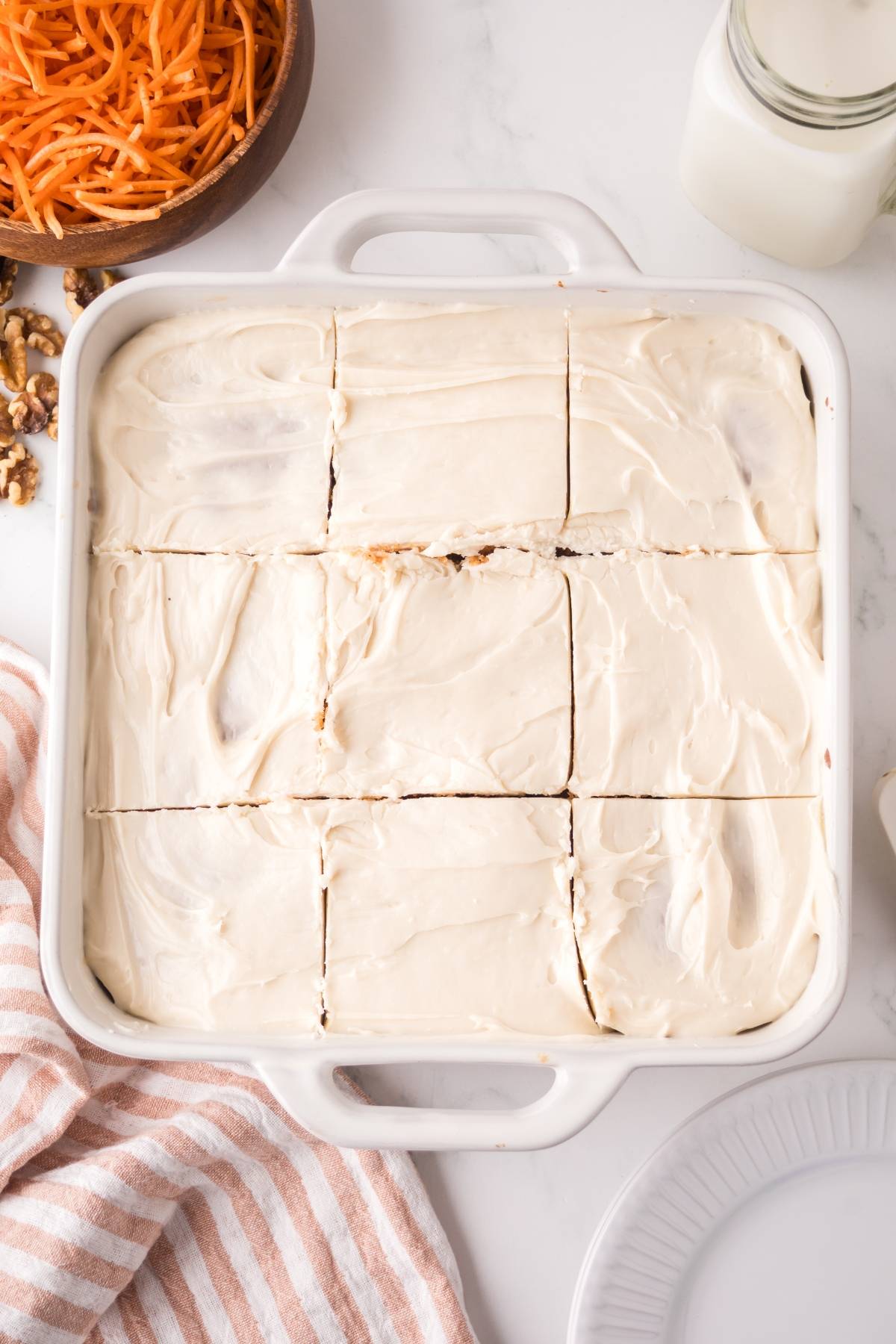 A square baking dish of frosted cake cut into nine pieces, with carrots and walnuts nearby.