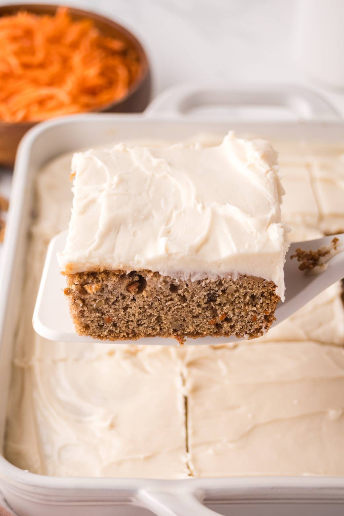 A piece of carrot cake with cream cheese frosting being lifted from a frosted cake in a baking dish.
