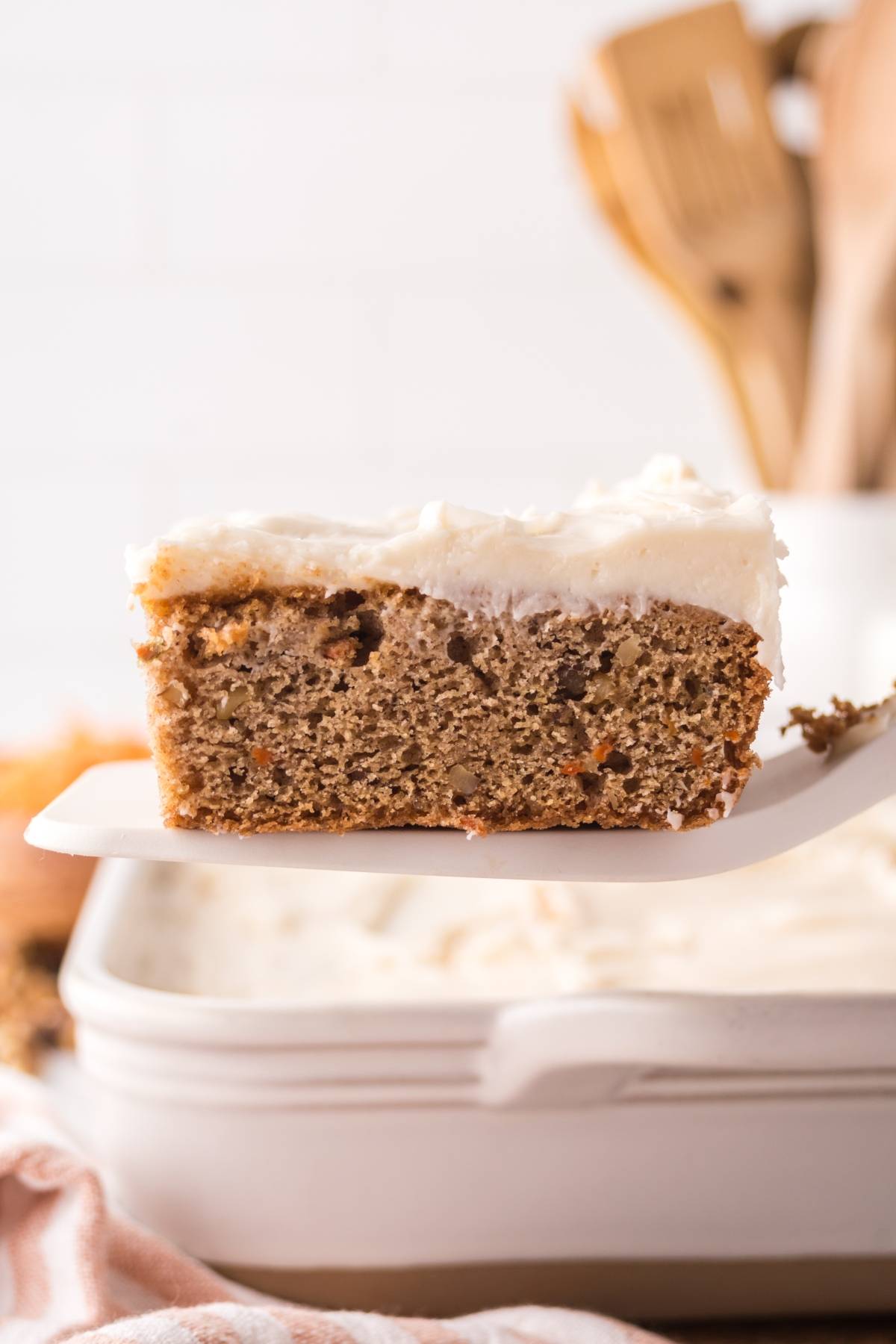 A slice of frosted banana cake on a spatula above a white baking dish.
