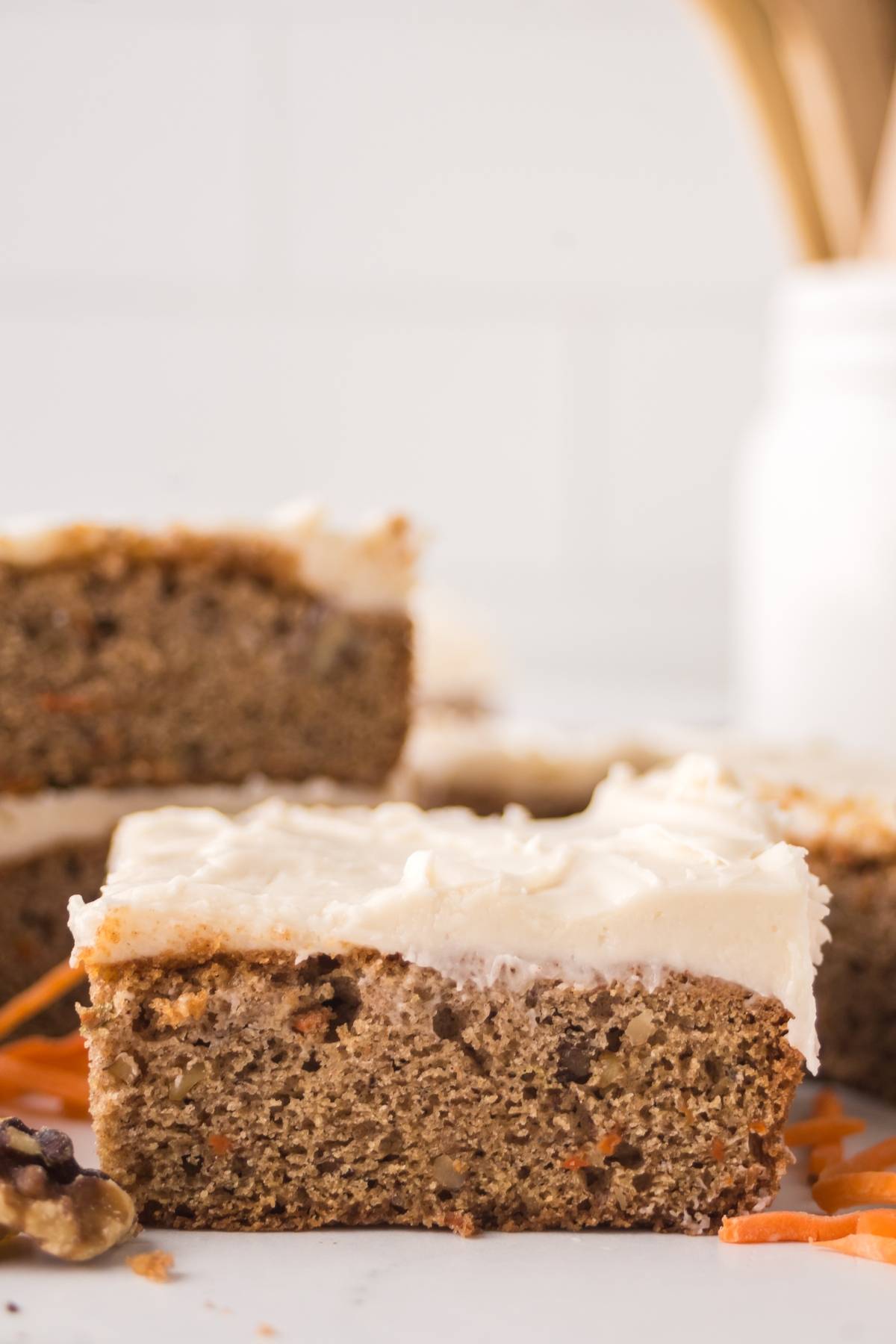 A close-up of a frosted carrot cake bar with a bite taken out of it, on a white surface.