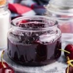 A jar of cherry jam is surrounded by fresh cherries and glass jars on a marble surface.