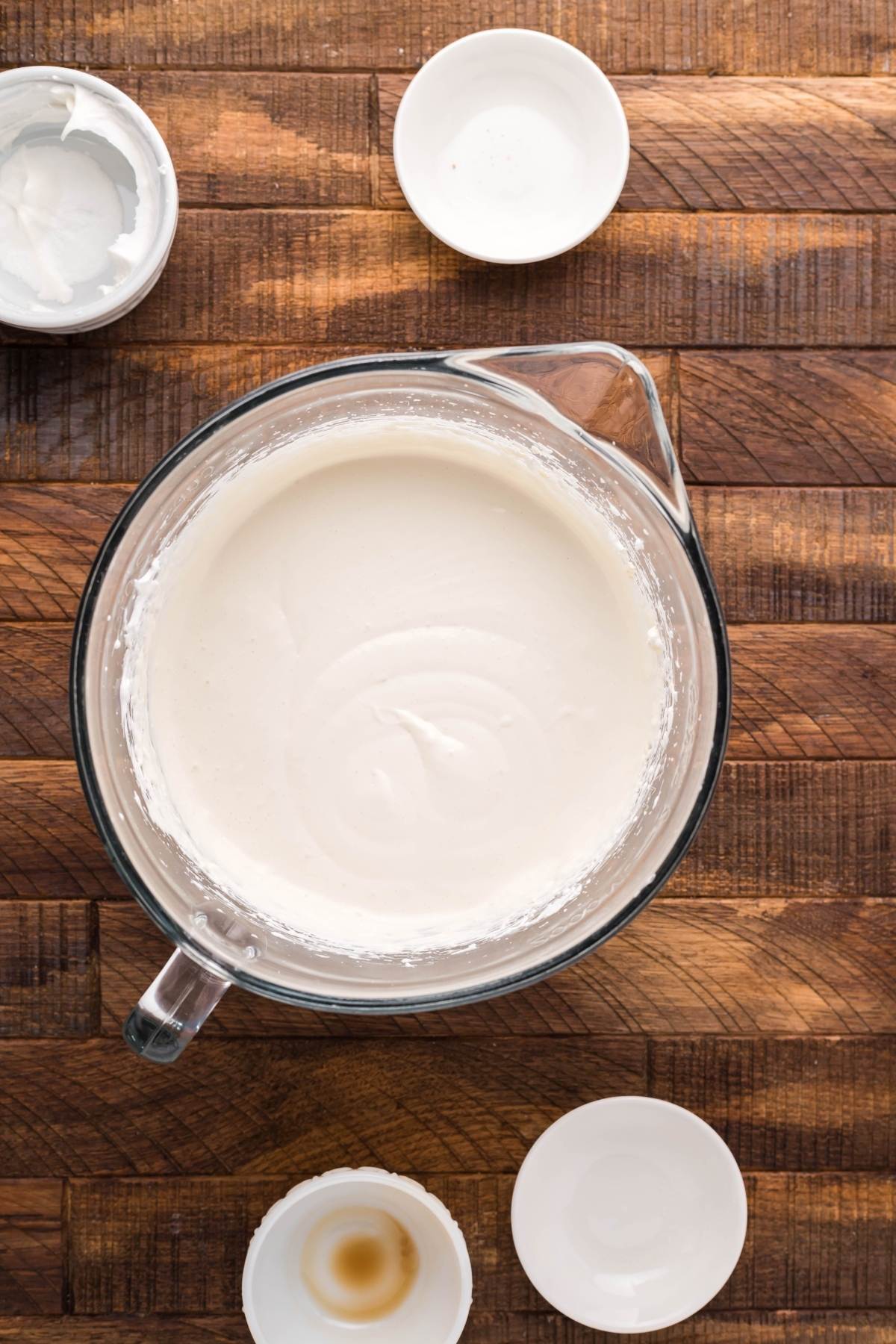 A glass bowl of whipped cream with four small empty bowls on a wooden surface, ready for assembling mini cheesecakes or cherry tarts with puff pastry.