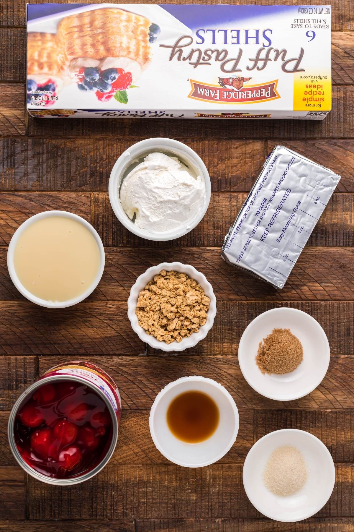 Ingredients for a dessert, including pre-made puff pastry shells, cream cheese, whipped cream, and bowls of various fillings—perfect for making mini cheesecakes or Cherry Cream Cheese Puff Pastries.