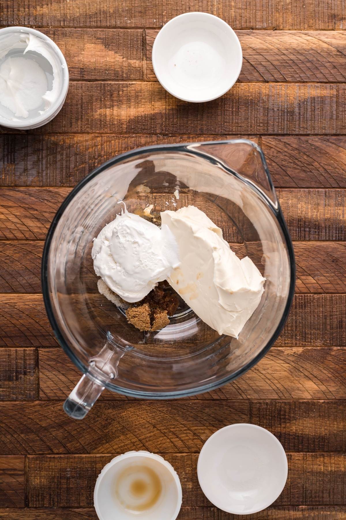 A glass mixing bowl with cream cheese, sour cream, and brown sugar on a wooden surface—an inviting start for mini cheesecakes—surrounded by small bowls.