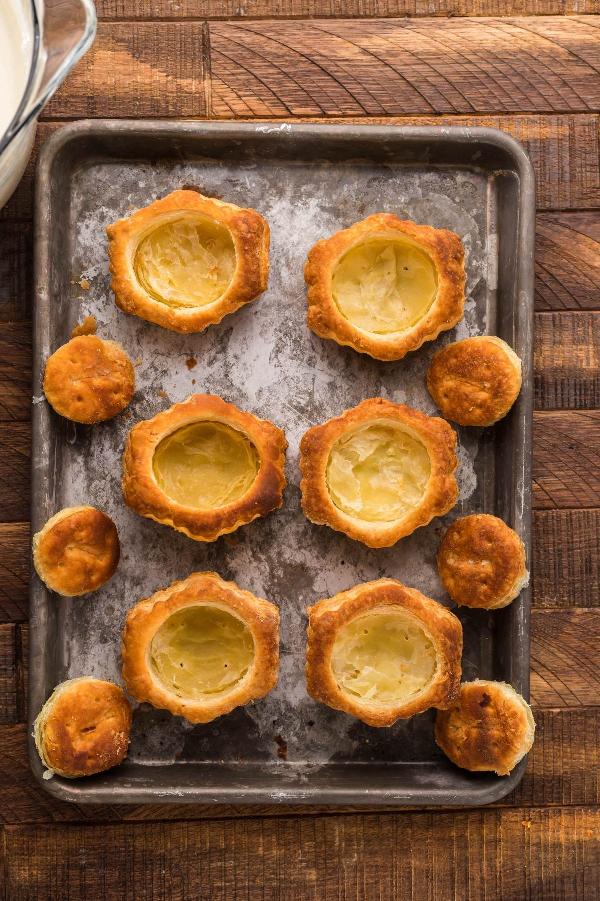 Golden pre-made puff pastry shells and lids rest on a baking tray atop a rustic wooden surface, perfect for mini cheesecakes or cherry tarts with puff pastry.