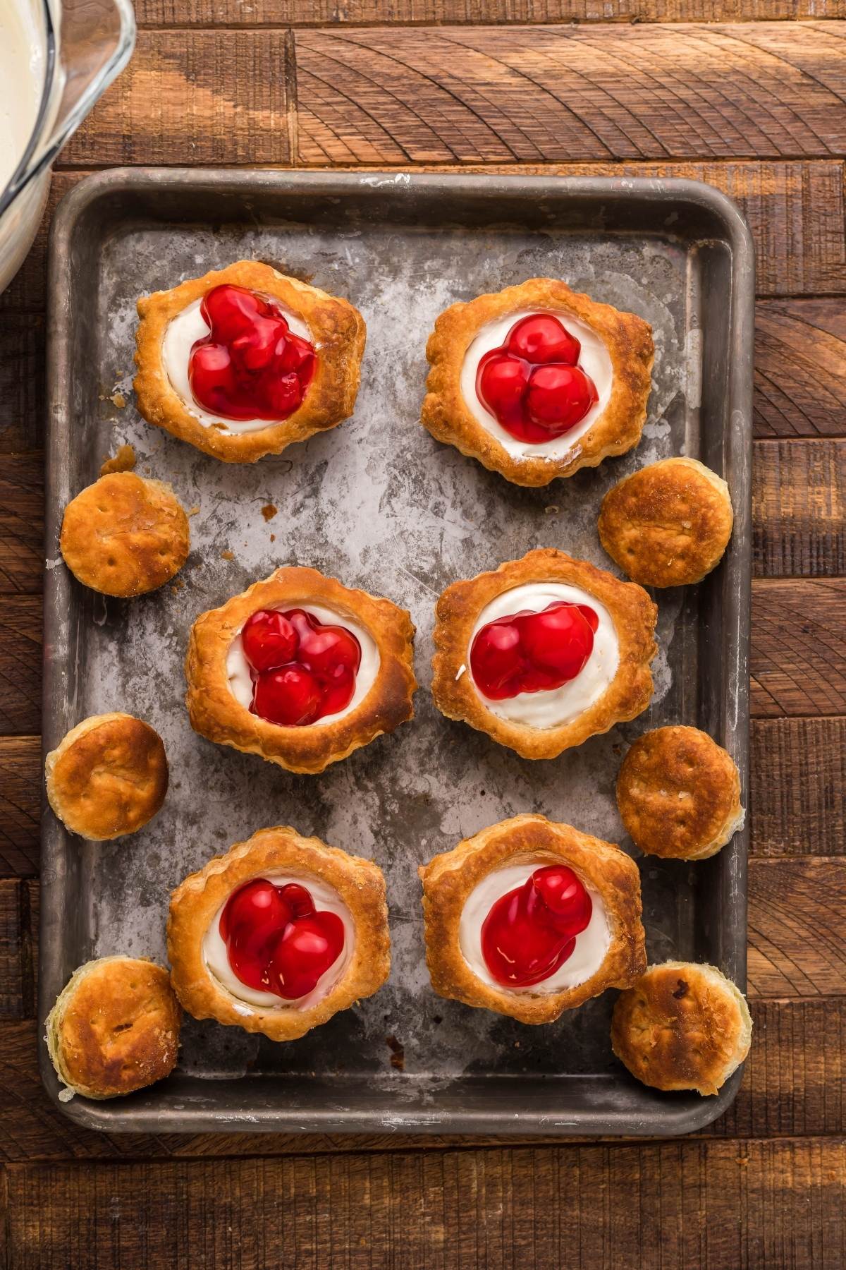 Six Cherry Cream Cheese Puff Pastries, each topped with a cherry and filled with cream, rest on a metal baking sheet atop a wooden table.
