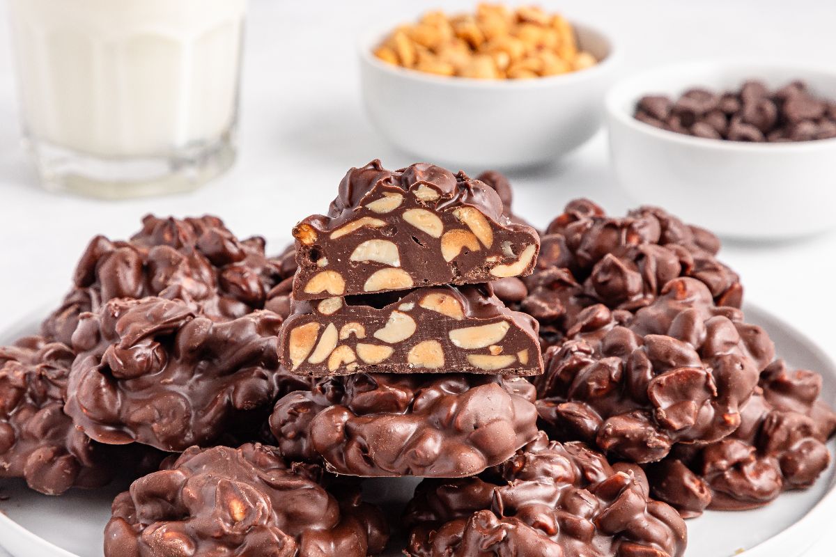 Crockpot Candy chocolate clusters with visible peanuts are stacked on a plate, surrounded by bowls of peanuts, chocolate, and a glass of milk in the background.