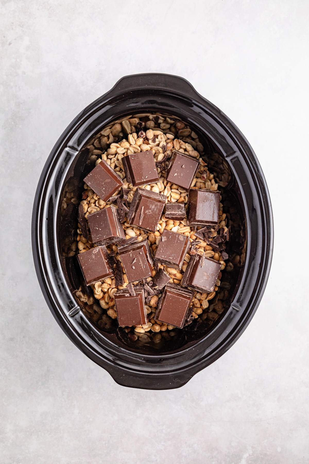 A slow cooker filled with oats, nuts, and square pieces of chocolate on top.