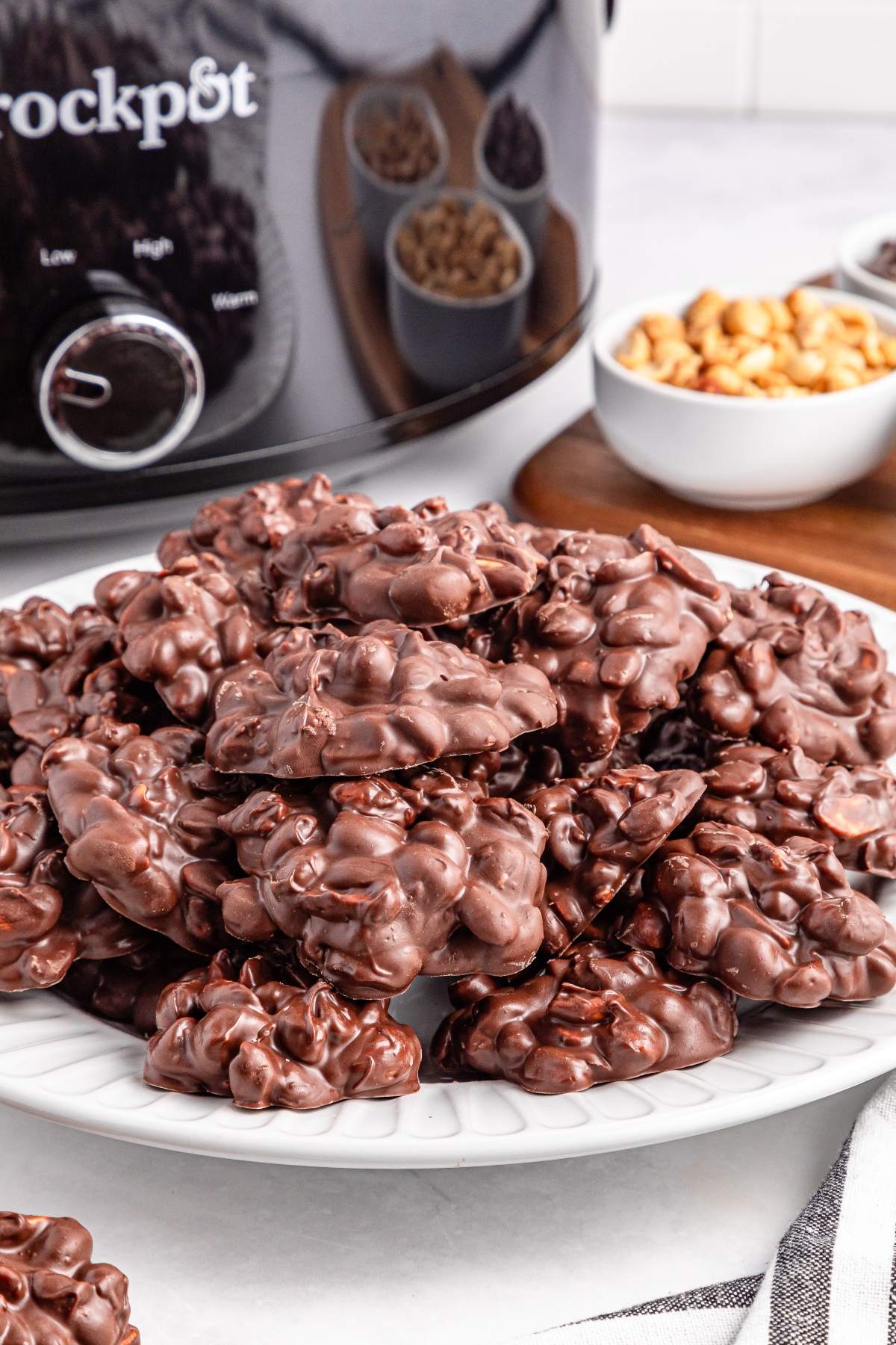 A plate of chocolate peanut clusters sits in front of a crockpot and bowls of peanuts and chocolate chips.
