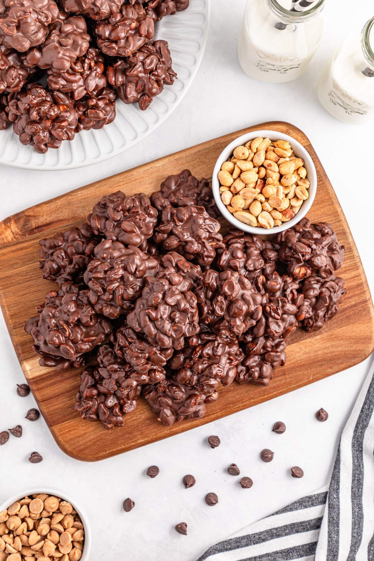 Chocolate clusters on a wooden board with bowls of peanuts, butterscotch chips, and bottles of milk nearby.