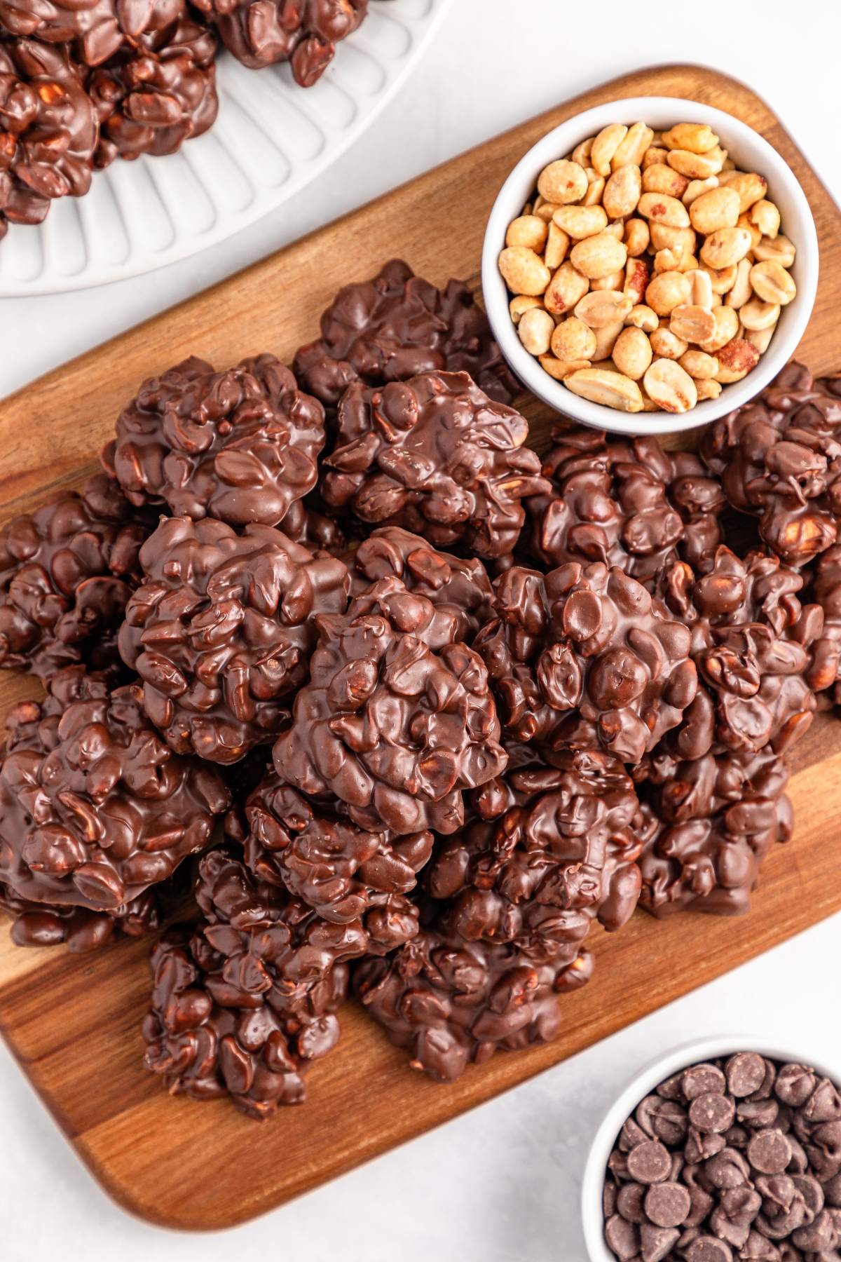 Clusters of chocolate-covered peanuts on a wooden board, with bowls of peanuts and chocolate chips beside them.