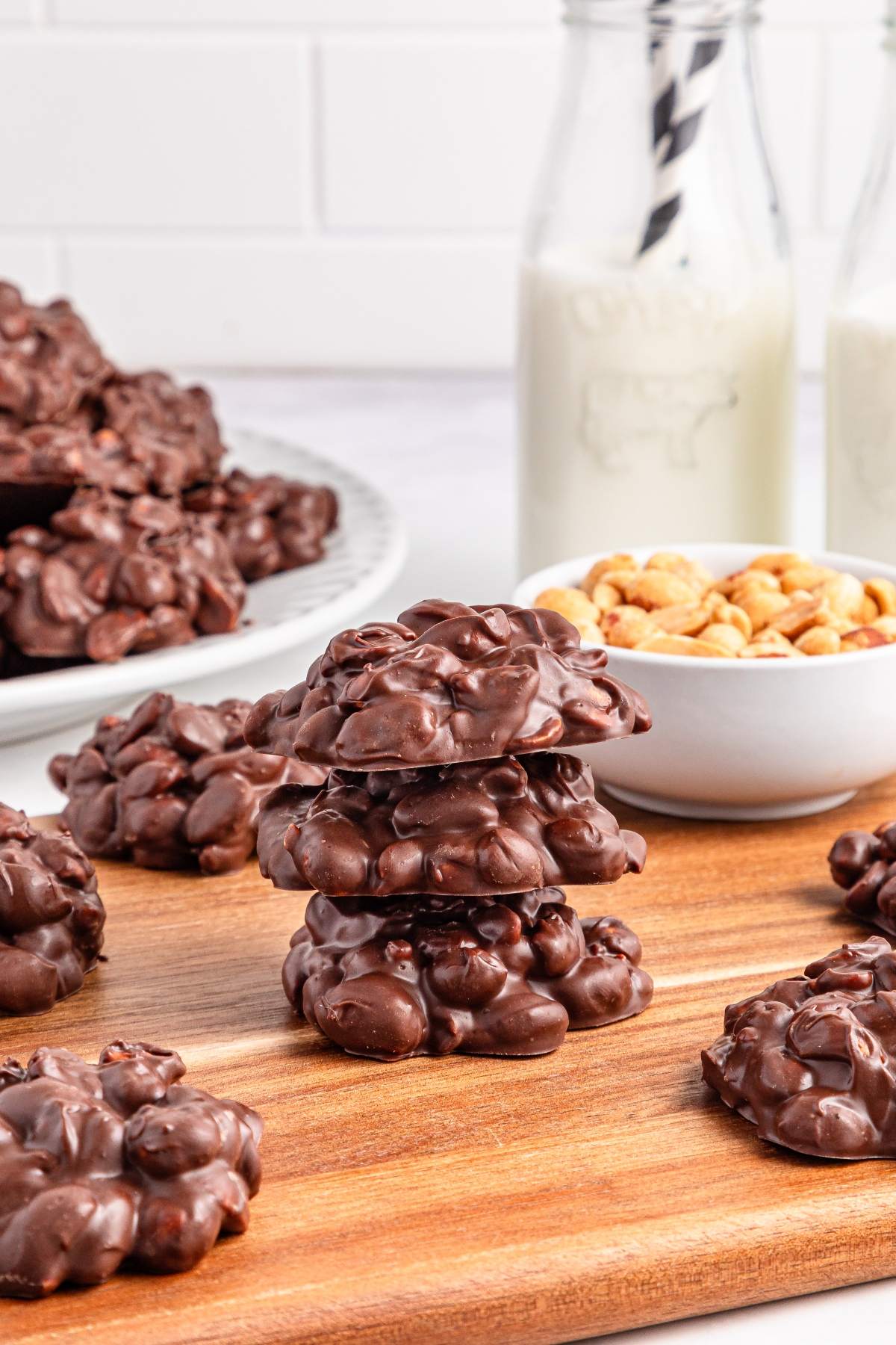 Chocolate peanut clusters stacked on a wooden board, with milk and peanuts in the background.