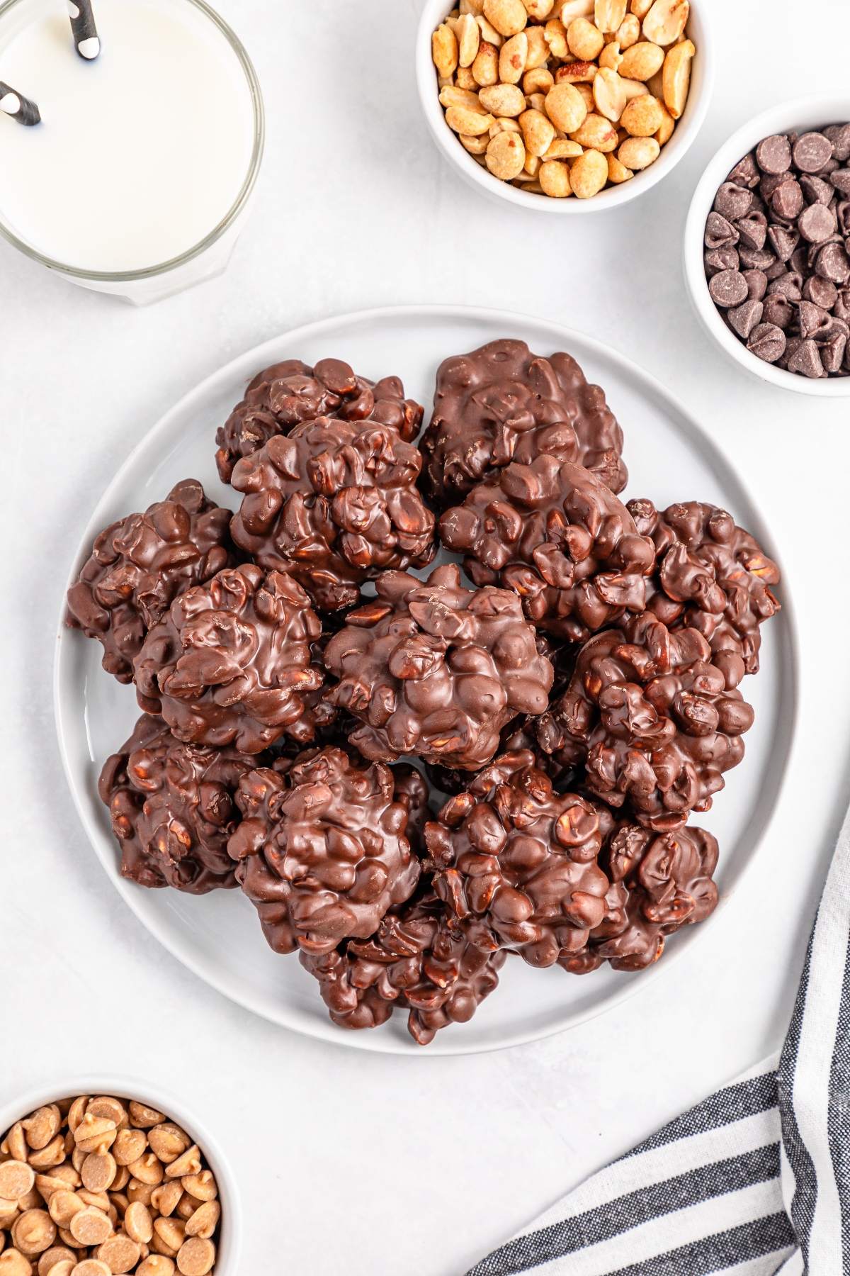 A plate of chocolate clusters with bowls of peanuts, chocolate chips, and a glass of milk nearby.