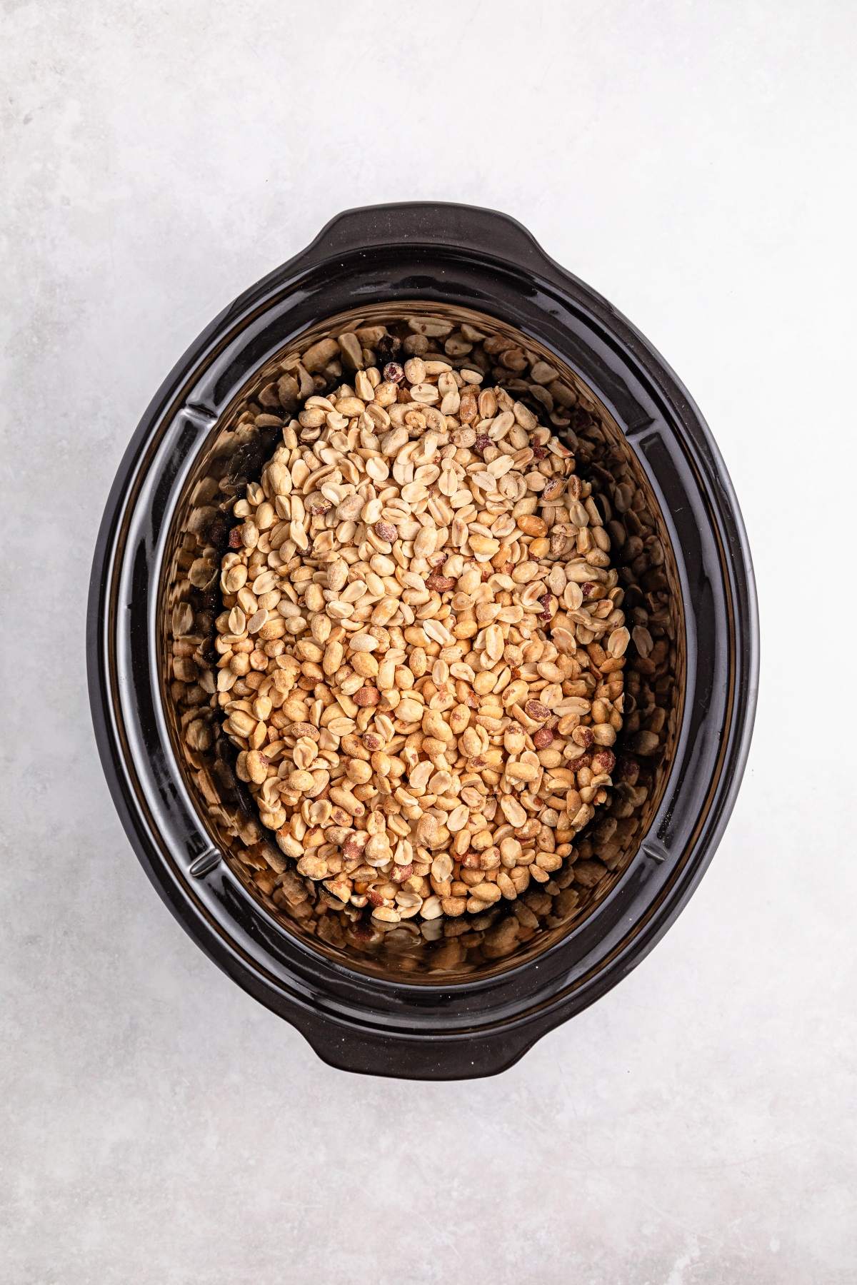 A slow cooker filled with raw, shelled peanuts on a light gray surface.