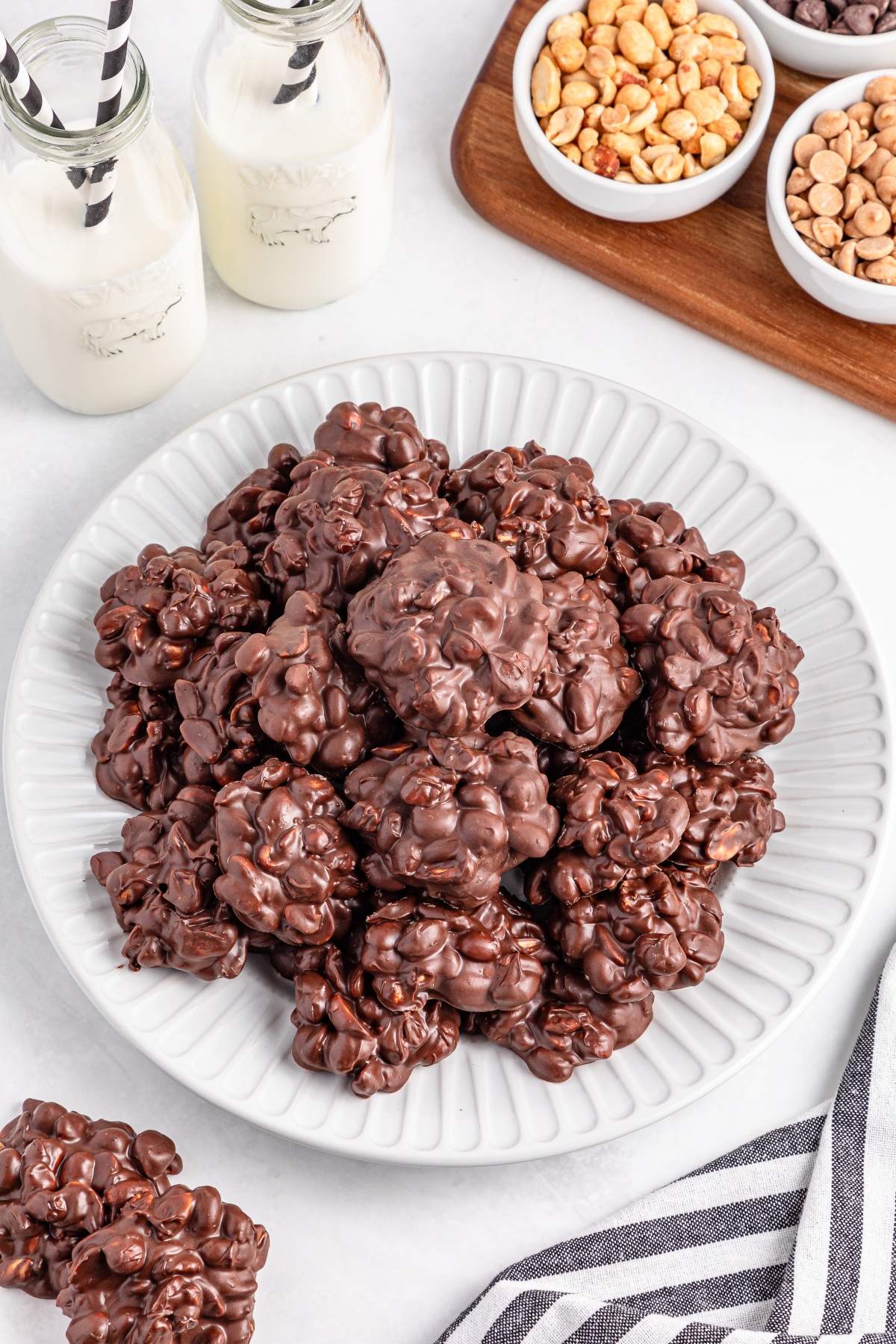 A plate of chocolate clusters sits next to bottles of milk and bowls of nuts and butterscotch chips.