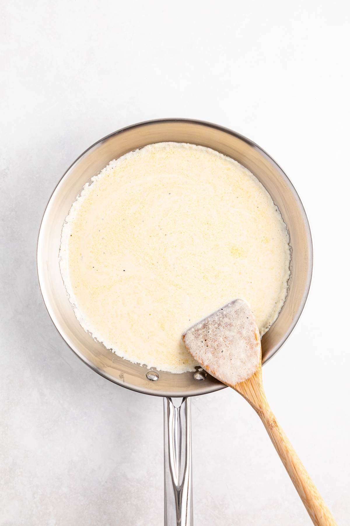 A stainless steel pan with a creamy sauce and a wooden spatula on a light surface.