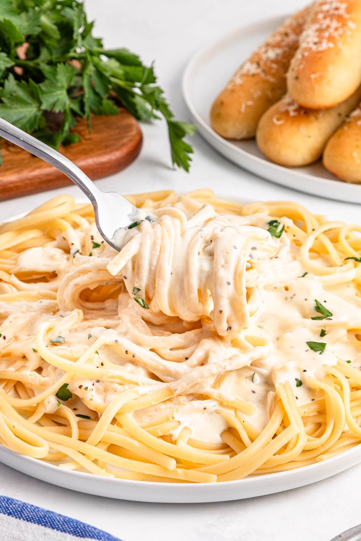 Fork twirls creamy fettuccine Alfredo on a plate, with breadsticks and parsley in the background.