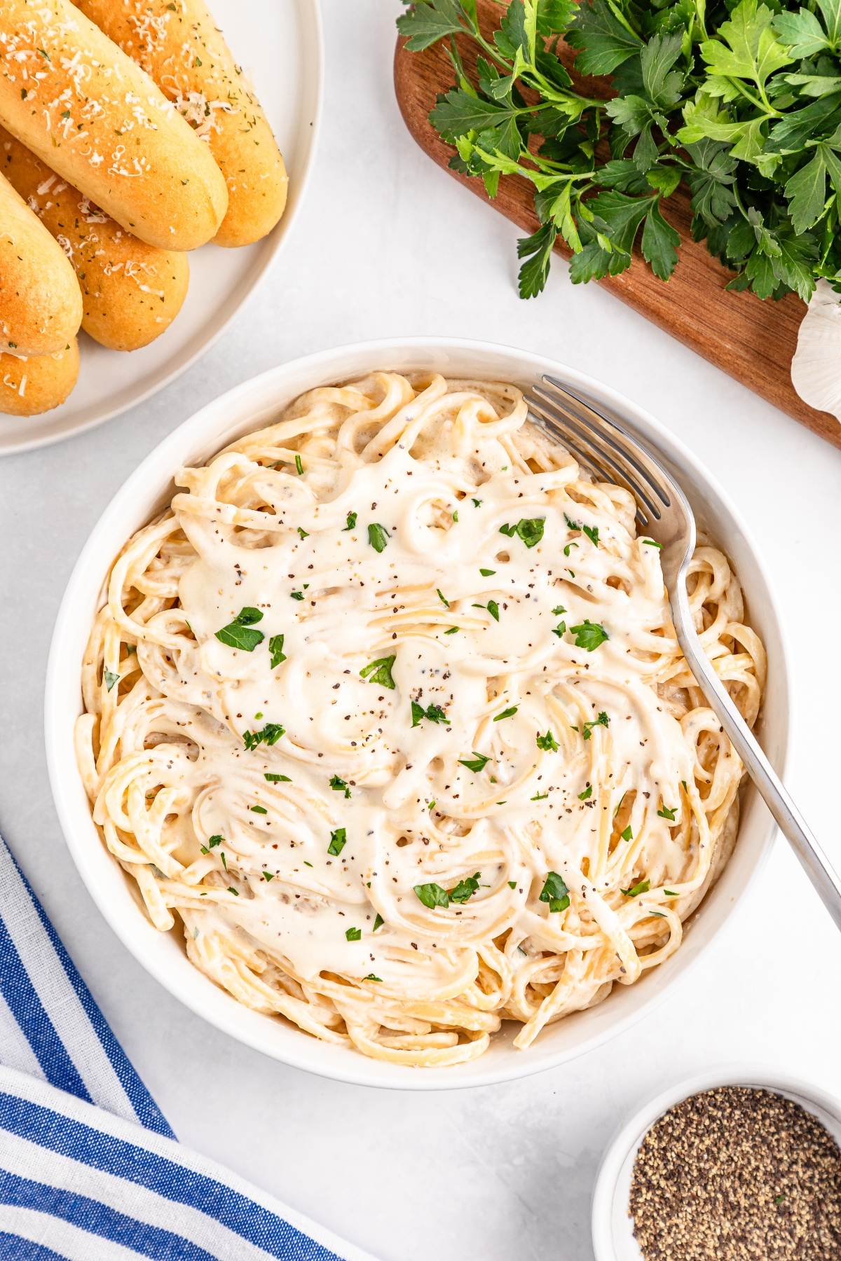 Bowl of creamy fettuccine Alfredo topped with parsley, next to breadsticks and fresh herbs on a white table.